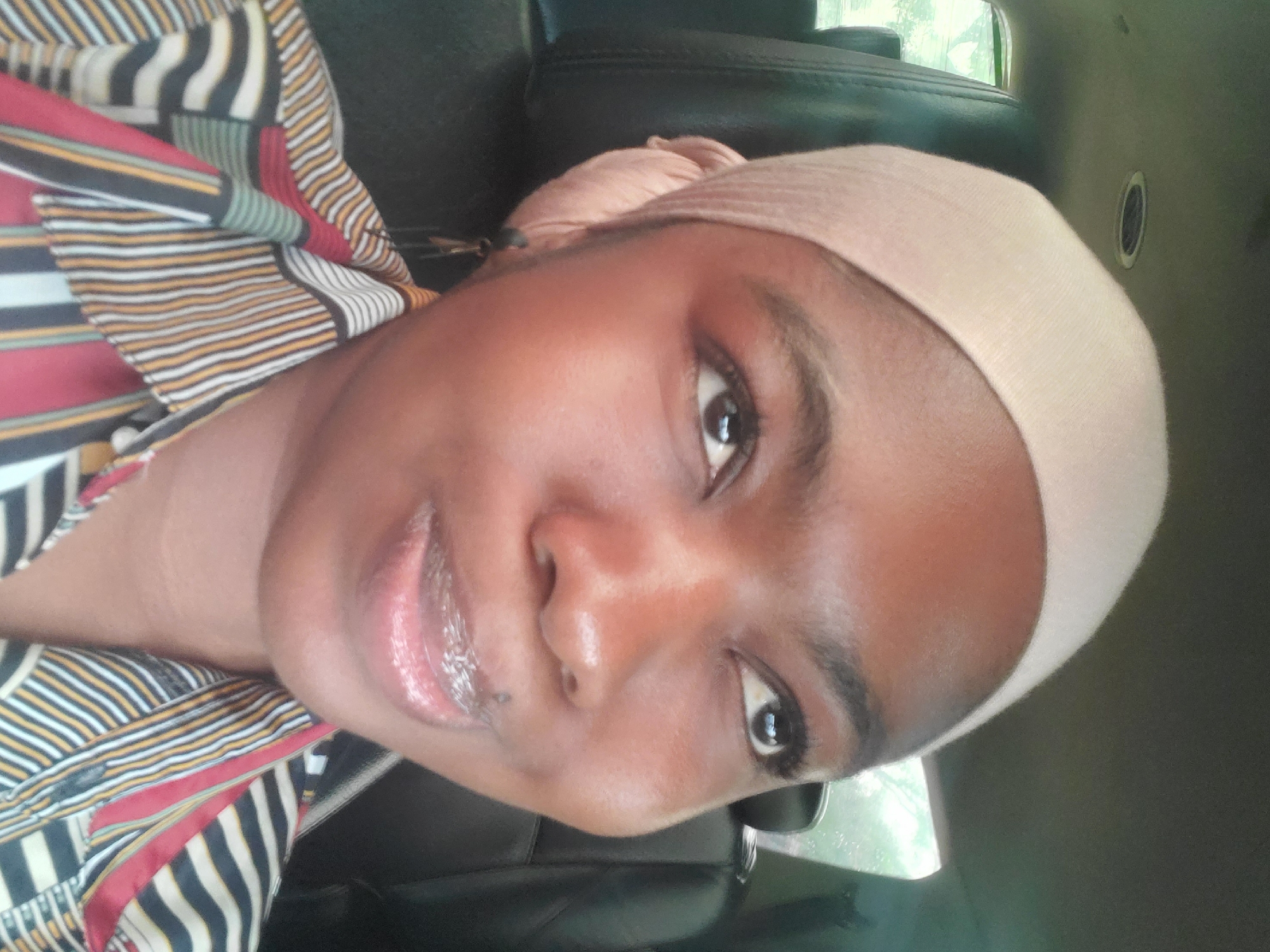 A close-up selfie of a dark-skinned woman with brown eyes. She's wearing a beige head covering and a patterned shirt with stripes and red accents. The background suggests she is inside a vehicle, possibly a car, with black leather seats. There's no information available to place this specific scene in Laima, Nigeria.