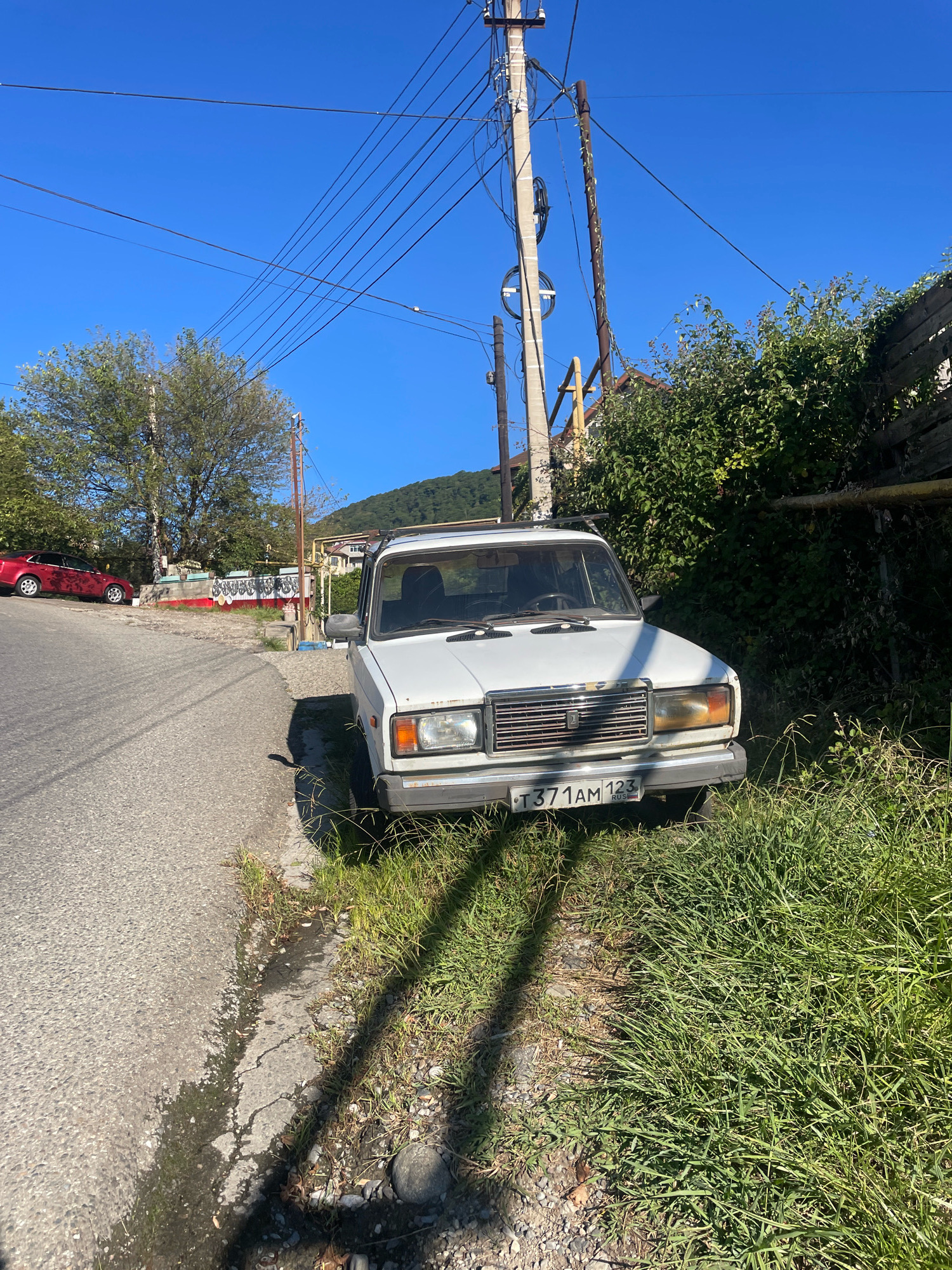 A white sedan is parked on the grassy shoulder of a paved road in Baranovka, Russia. The car, a Lada or similar Soviet-era model, faces the camera and is angled slightly towards the right. Its license plate reads "T371AM 123 RUS". The road curves to the left in the foreground, and a red car is visible further up the road on the left. Lush green vegetation, including trees and bushes, surrounds the scene. Overhead, a tangle of power lines crisscrosses the bright blue sky, supported by utility poles. The environment appears to be a rural or suburban area, with a hilly landscape visible in the background. Sunlight casts long shadows across the road and grass. There are no people visible in the image.