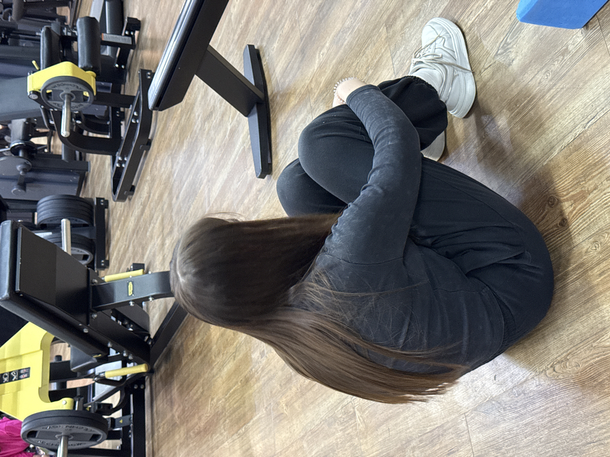 A woman with long brown hair is seated on a wooden floor in a fitness facility in Chaikovsky, Russia. She is wearing black pants and a long-sleeved black shirt and white sneakers. Her legs are drawn up to her chest, and she is hugging her legs. Fitness equipment, including weight machines, is visible in the background.