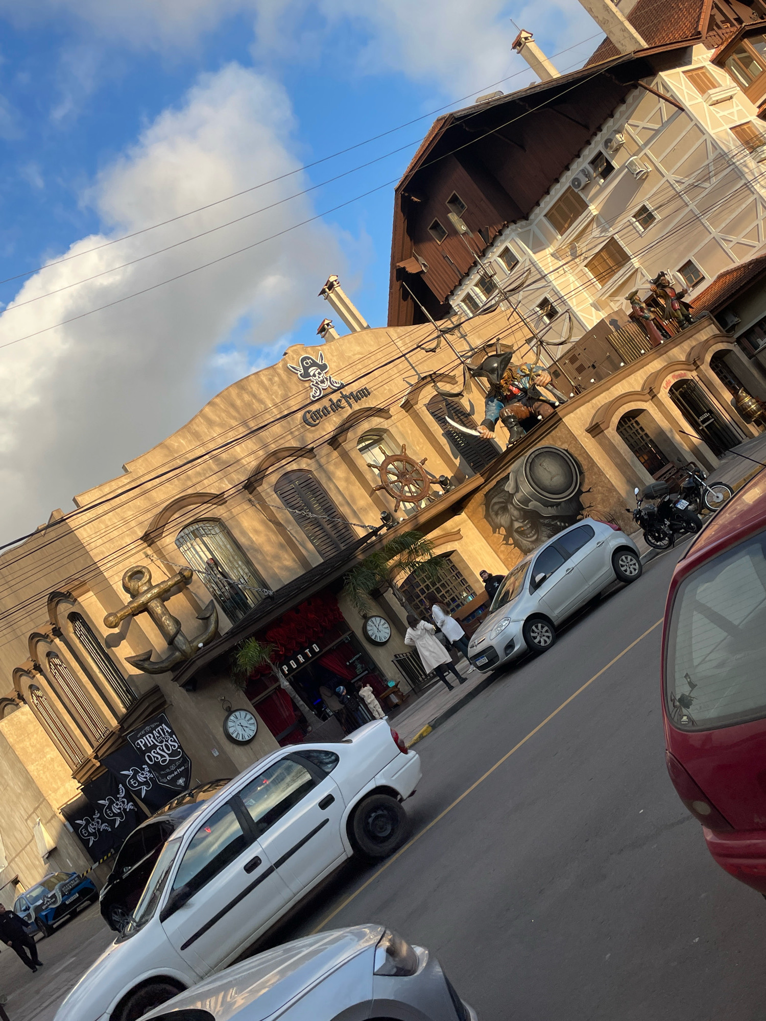 The image shows a street scene in Gramado, Brazil, on a partly cloudy day. A building with a pirate-themed facade, possibly a restaurant or attraction, is prominently featured. The building has several decorative elements such as a large anchor, a ship's wheel, and figures of pirates. The name "Casa de Brio" is visible on the facade, along with a pirate logo. A silver car is parked in front of the building, and a red car is partially visible in the foreground. Several people are seen walking on the sidewalk. The street appears to be paved, and there are power lines overhead. The architecture of the building suggests a European influence, which is common in Gramado.