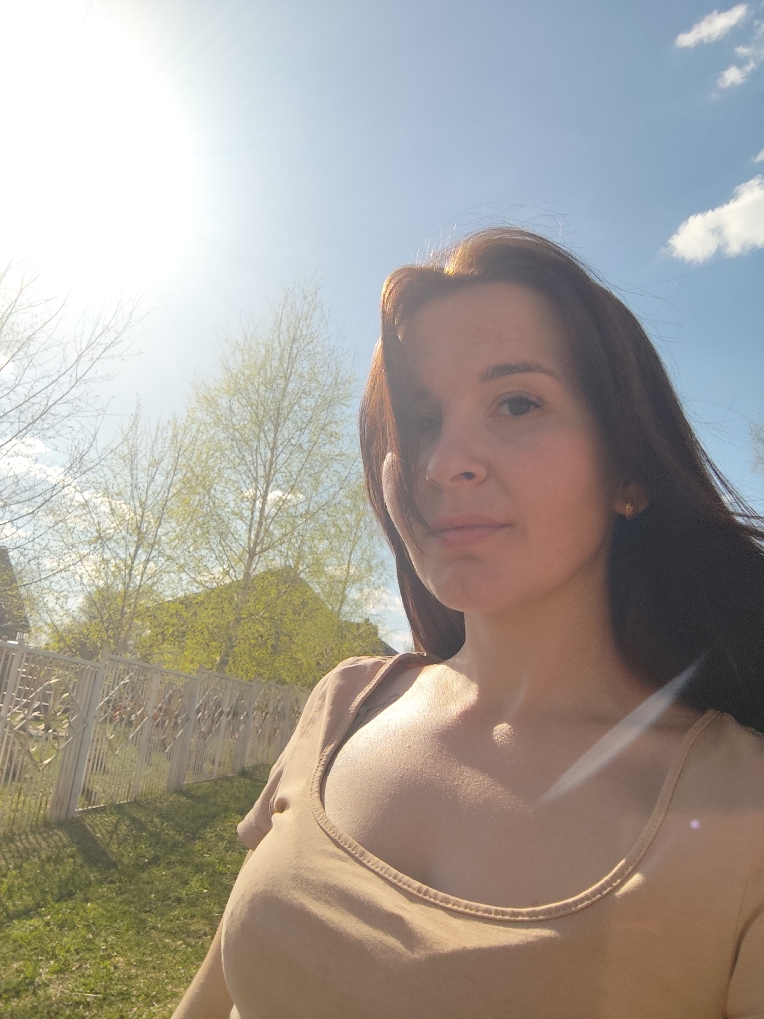 A woman with dark brown hair stands outdoors, looking towards the viewer, likely in a self-portrait style. She is wearing a light brown top. The background includes a bright, sunny sky with a few clouds, a fence, green grass, and trees. Based on the prompt information, the location is Фоки, Russia.
