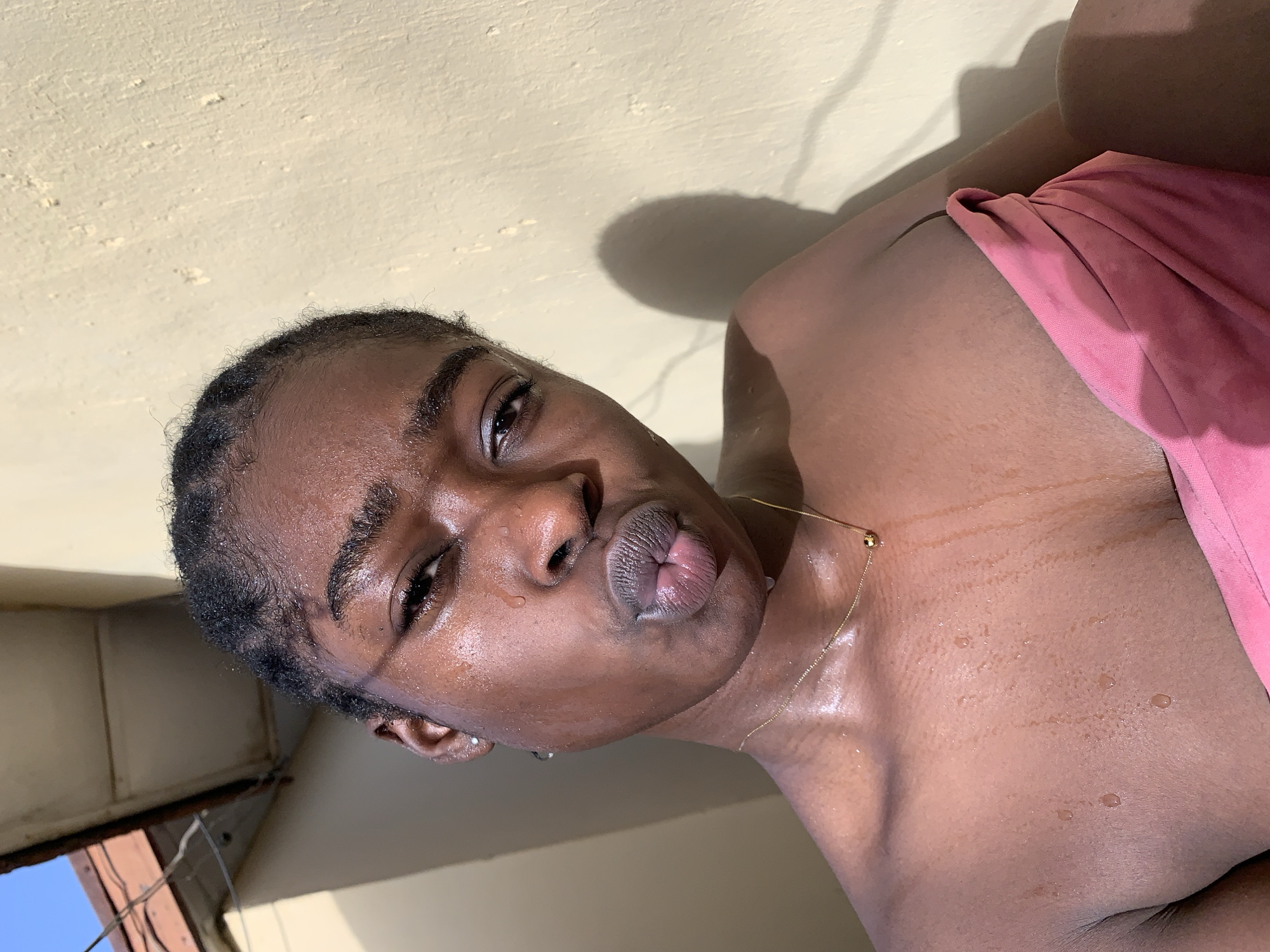 A close-up, indoor shot depicts a dark-skinned person with short hair. The person is slightly angled, facing the camera. They have wet skin. A thin gold necklace is visible. Part of a pink fabric is visible, draping over the person's upper body. The background is a beige wall. Cables and a blue sky are visible through a window. The image appears to be taken from a low angle. The location in the city of Rafin Par, Nigeria, cannot be determined.