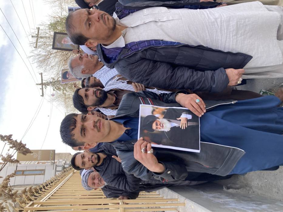 A group of approximately ten men are depicted in a daylight outdoor scene within Quetta City Tehsil, Pakistan, participating in what appears to be a procession or demonstration. The sky is overcast.
In the foreground, a young adult male, estimated to be in his 20s, with a mustache, wears a black leather jacket with red and white stripes on the shoulders over a blue traditional garment. He holds a laminated portrait of an elderly bearded man in a black turban and robe, identified as a religious leader, with his right thumb obscuring a small portion of the image. He wears a red ring on his right ring finger and a turquoise ring on his left ring finger. His expression is serious, and he looks slightly to his right.
To his immediate right, a middle-aged male wears a light gray traditional shalwar kameez under an open black puffer jacket with a visible blue patterned lining. He has a stern expression, looking directly forward, and wears a silver ring on his left ring finger.
Behind these two individuals, other men are visible in a line. One man in a black puffer jacket and a dark beard stands directly behind the central figure. Further back, an older man with a white turban is present. To the right, behind the middle-aged man, a male with a dark beard wears a brown garment and a white and black checkered scarf around his neck. An older male with short gray hair, wearing a plaid shirt, is seen holding a framed portrait of a religious figure. Another similar framed portrait is visible further in the background.
The immediate left background features a tall, dark metal fence or gate with vertical bars. Behind the fence, a light-colored building with a sign displaying "MAZGI" is partially visible. The distant background shows power lines, utility poles, and bare tree branches against the cloudy sky.