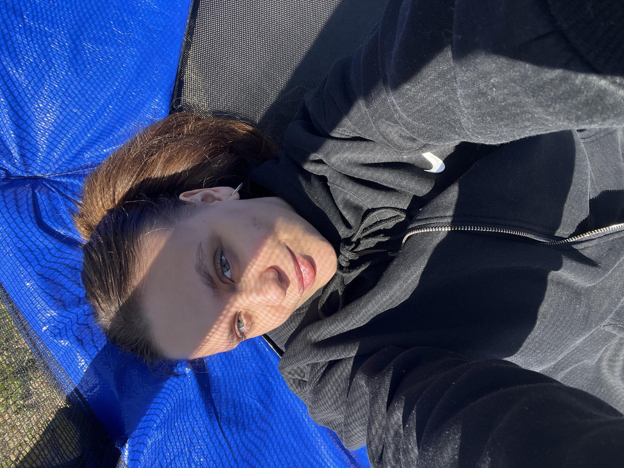 A fair-skinned person with light eyes and brown hair pulled back in a ponytail lies on a blue and black surface, likely a trampoline, in an outdoor setting. The person is wearing a black, zippered hoodie. Sunlight casts striped shadows across their face. The setting appears to be outdoors, possibly a yard or recreational area, evidenced by the grass visible in the lower left corner. The photo is taken from a close-up, slightly angled perspective, looking down at the person. There is no definitive indication of this photo being taken in the city of Ольховка, Russia.