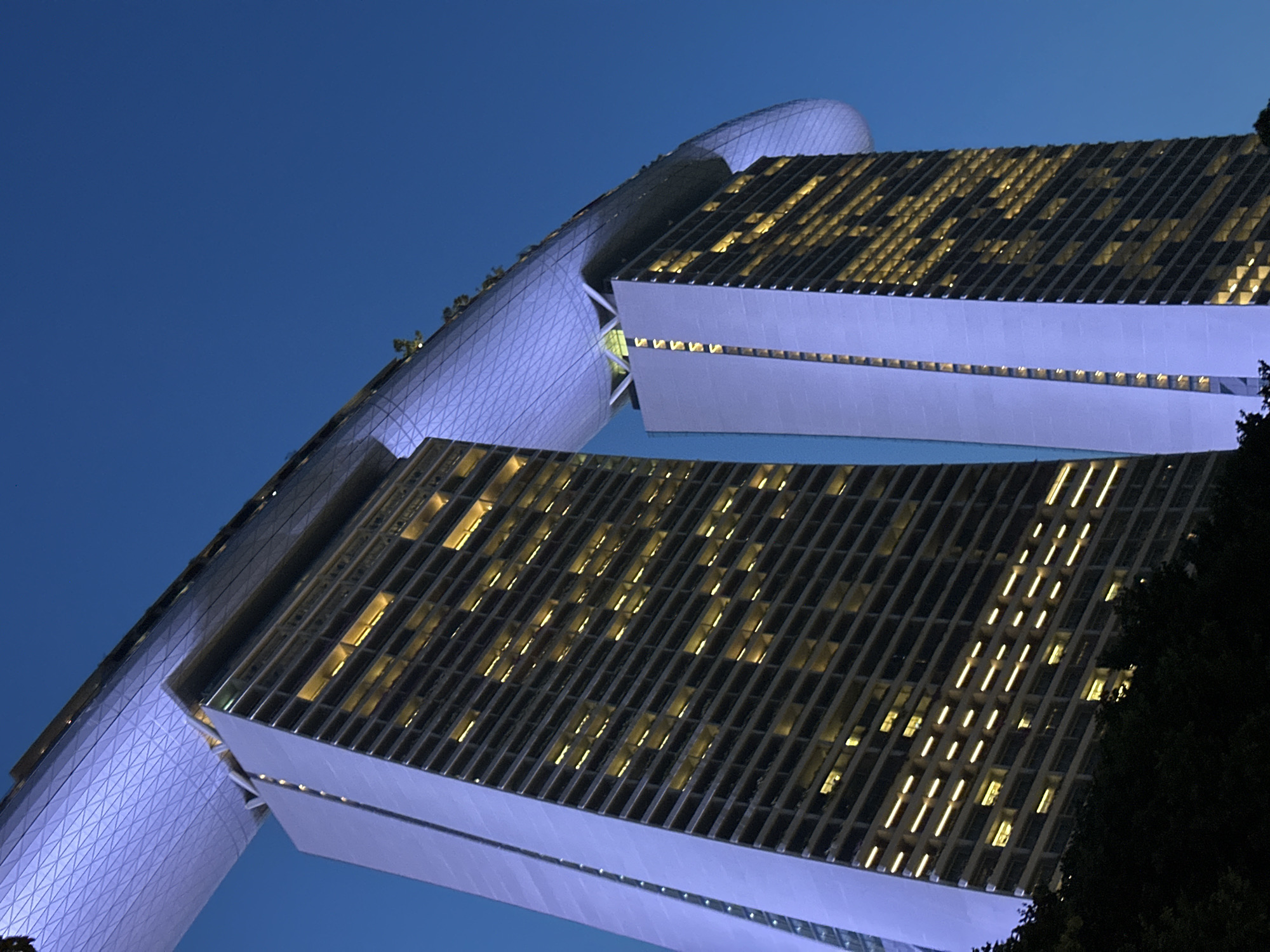 The image shows a low-angle, wide shot of the Marina Bay Sands hotel in Singapore at dusk. The iconic three-tiered skyscraper complex features two of its three towers prominently in the frame, with the connecting SkyPark visible on top. The towers are illuminated by a cool blue light on their sides, contrasting with the warm yellow glow of the windows from within. The SkyPark, shaped like a ship, has a complex geometric patterned roof illuminated by a soft purple light. There are trees in the foreground, silhouetted against the buildings, suggesting a park or garden area. The sky is a deep, clear blue, indicative of twilight. No people or vehicles are visible.