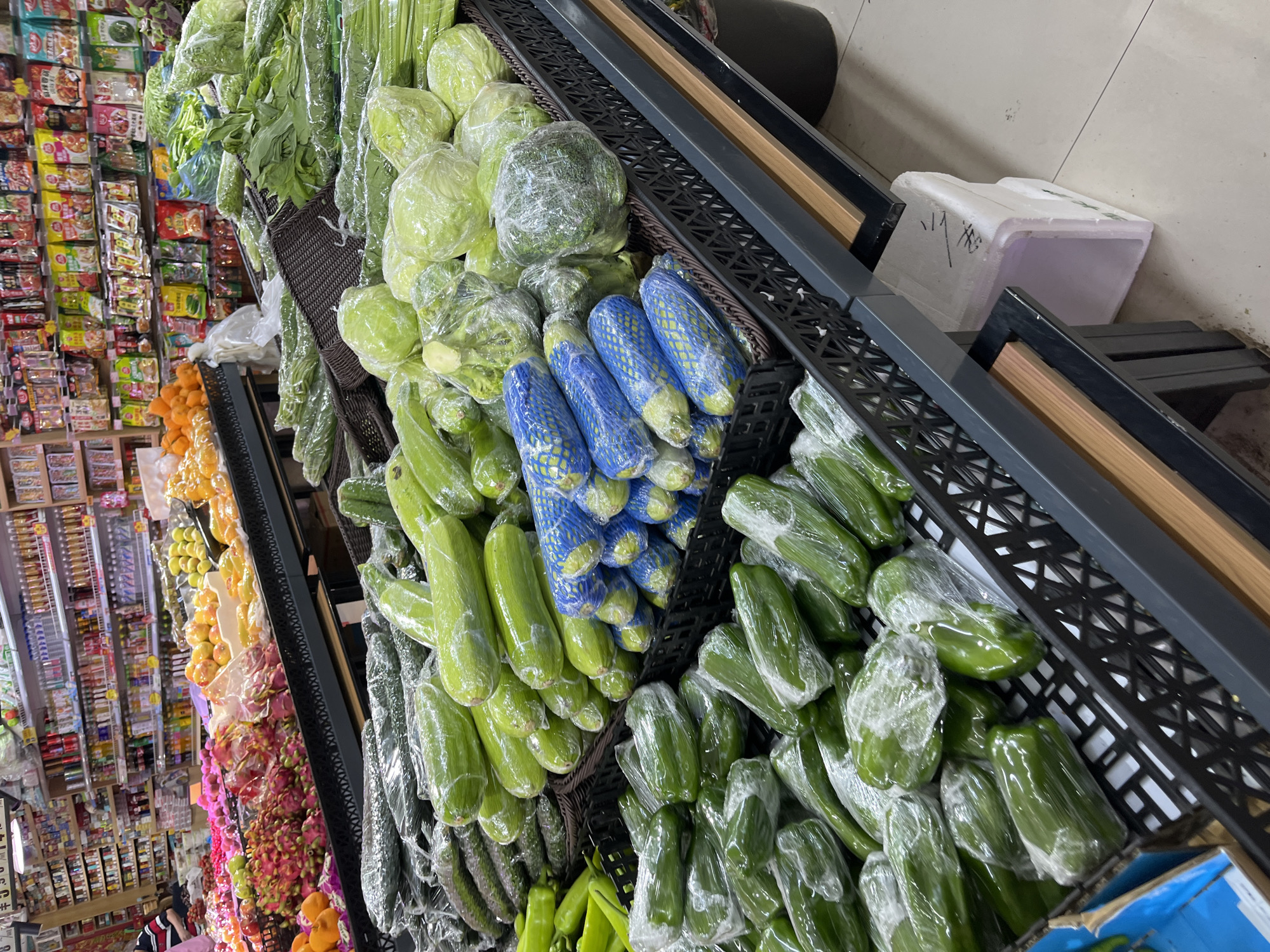 The image shows a grocery store shelf in Changchun City, China, stocked with fresh produce. A variety of vegetables, including wrapped cucumbers, zucchini (some in blue mesh), cabbage, broccoli, and celery, are displayed in black wire baskets. Behind the produce are shelves filled with packaged goods, including various types of instant noodles. In the background, there is a display of fruits and other packaged goods. The floor is tiled and there's a white container next to the produce display.