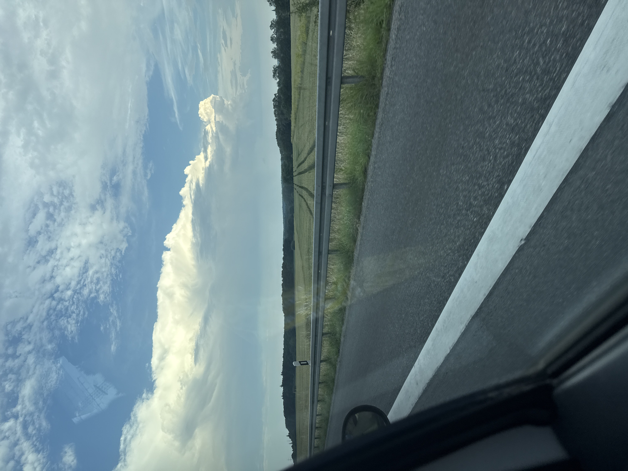 The image is taken from a moving vehicle on a highway in Linden, Germany. The foreground shows the asphalt road with a white line marking the lane. To the left, the interior of the car is visible, including a side mirror and a portion of the car door. In the midground, a metal guardrail separates the highway from a field of green crops. The field has visible tracks, suggesting recent agricultural activity. A single white and black road marker stands in the field. The background features a dense line of trees, forming a forest, under a sky filled with dramatic clouds. The clouds are a mix of bright white cumulus formations and darker, more scattered clouds, with patches of blue sky visible. The overall scene depicts a rural landscape seen from a highway.