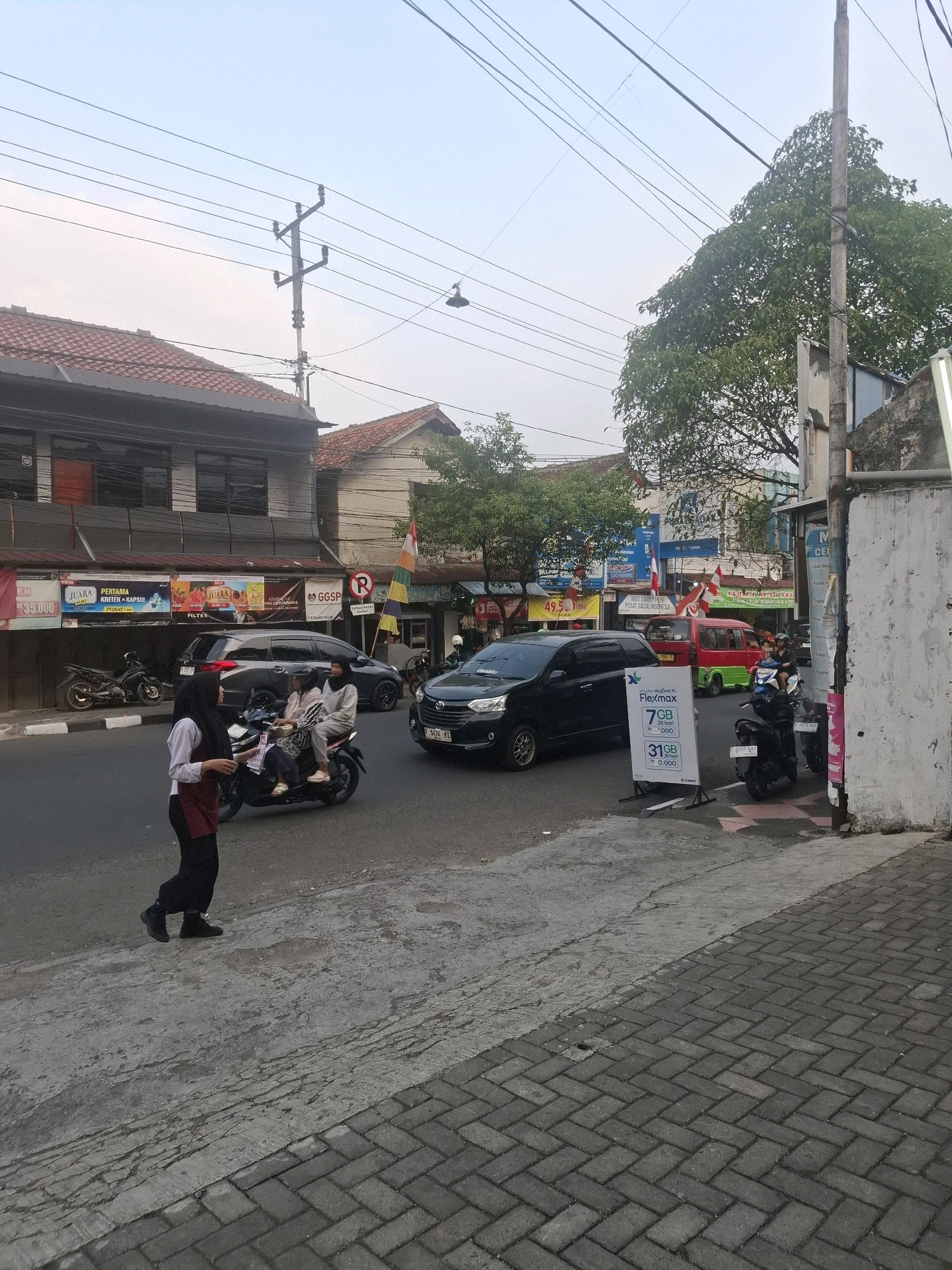 This is a street scene in Cianjur, Indonesia, with buildings lining the left side of a road. A sidewalk is visible in the foreground, constructed with gray interlocking bricks. A woman in a white shirt, red vest, and black pants is walking on the sidewalk away from the camera, facing left. In the street, a motorcycle carries two passengers. To the right of the motorcycle, a black car is visible. Further down the street, a red and green van is parked. A white stand with advertisements for data plans is placed on the sidewalk to the right of the street. Power lines crisscross the sky above. The buildings appear to be shops, with several signs visible, including one for "GGSP" and another that is a red and white flag. The sky is overcast.