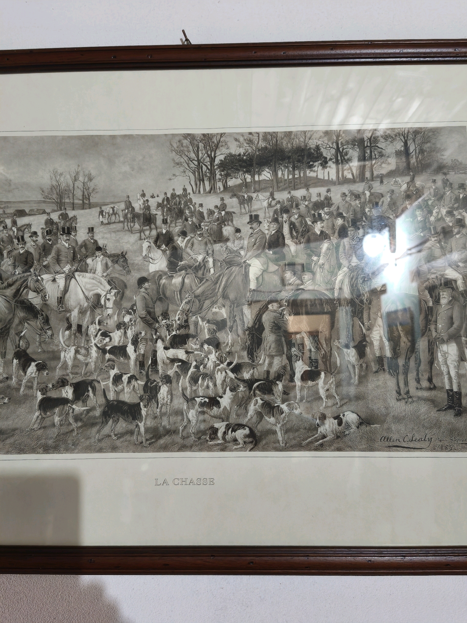 This is a framed print of a hunting scene. The print depicts a large group of people on horseback and a pack of hounds in a field with trees in the background. Some of the people are dressed in hunting attire. The scene is rendered in black and white. The title "LA CHASSE" is printed below the image. The artist's signature, "Allen C. Lealy," is visible in the bottom right corner of the print. The print is framed in a dark wood frame. The image was taken indoors, and there are reflections on the glass of the frame, including a bright white light source. There is no indication of the location in the city of Passignano sul Trasimeno, Italy.