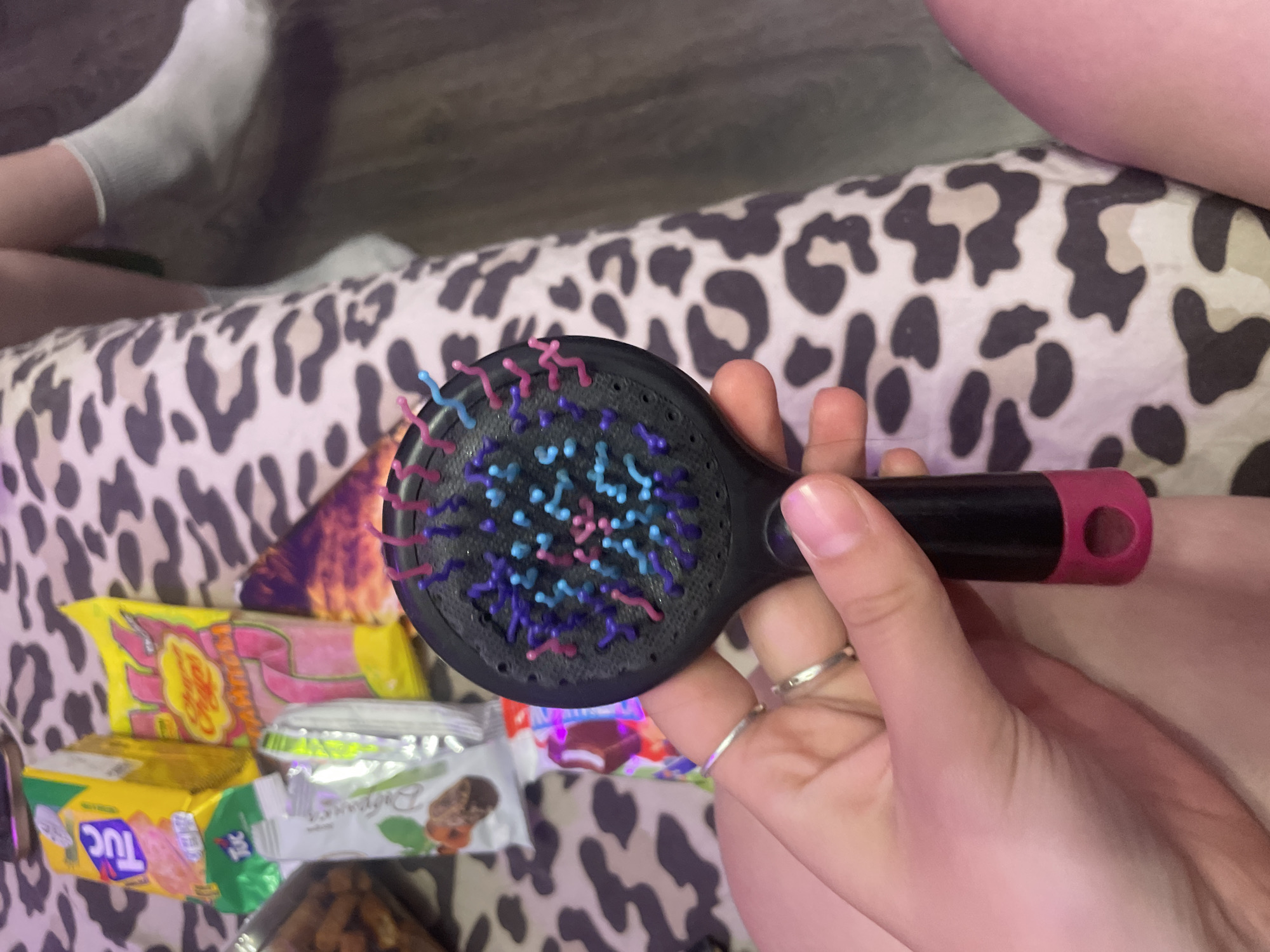 A close-up, eye-level shot shows a person's hand holding a black hairbrush with multi-colored bristles. The hand is positioned in the foreground, with fingers adorned with silver rings. The hairbrush is dark with purple, pink, and blue bristles. In the background, out of focus, is a surface covered with a leopard print fabric. To the left of the hairbrush, several candy wrappers are visible, including a yellow "TUC" cracker package and a pink "Chupa Chups" lollipop package. To the right of the hairbrush, a person's legs are visible, wearing grey socks and leaning against the leopard print surface. The overall lighting suggests an indoor setting. The image does not provide any information about the location in the city of Shcheglovo, Russia.
