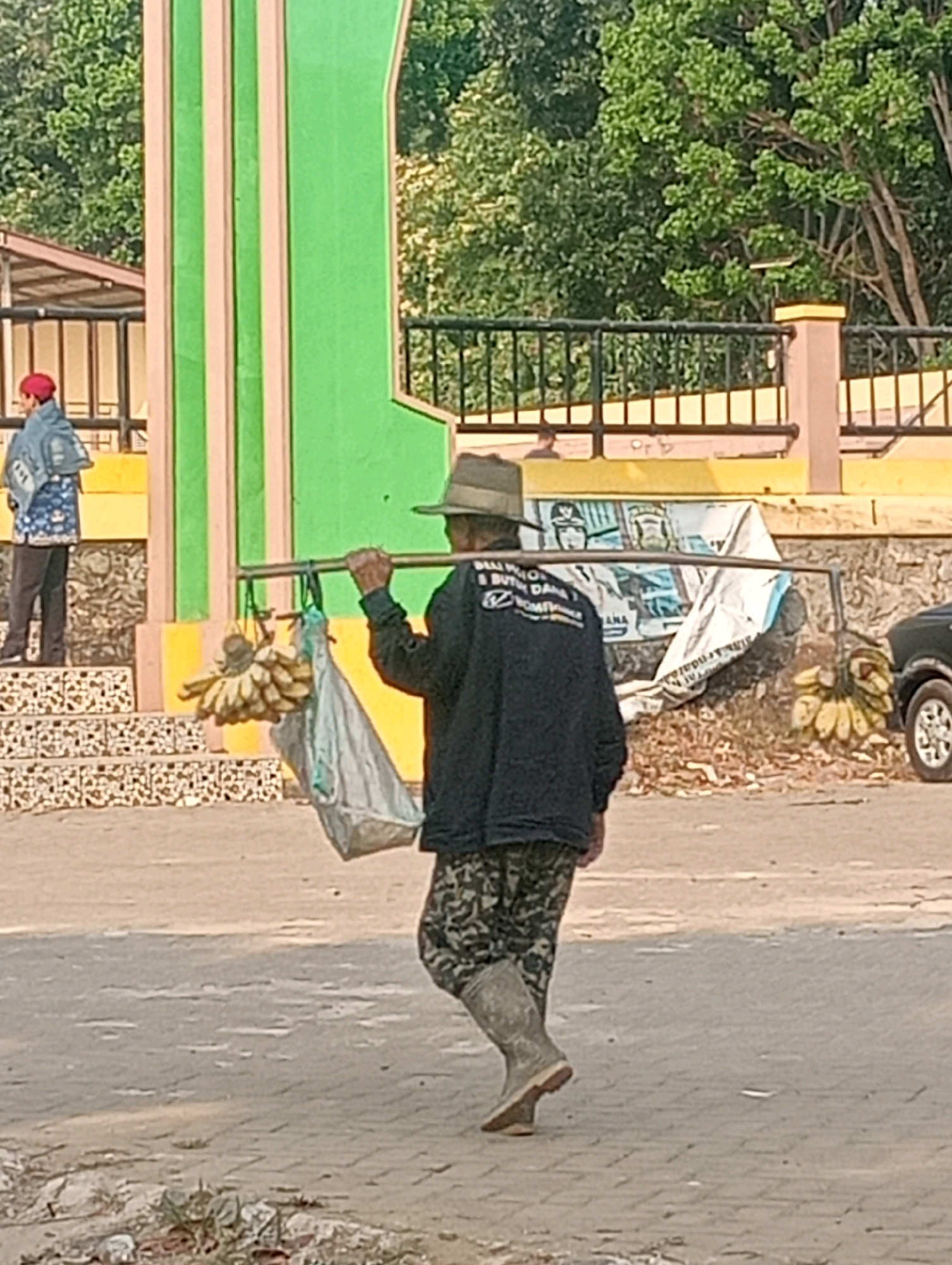 A man wearing a hat, a dark jacket, camouflage pants, and boots walks across a paved surface. He carries a long pole horizontally across his shoulders, with bunches of bananas hanging from one side and a large plastic bag from the other. In the background, a person wearing a red hat and a blue jacket with a pattern stands near a structure with green and tan vertical stripes. Beyond that, there is a metal fence and trees. The scene appears to be outdoors during the daytime, under natural light. The area has a tropical feel with lush green foliage. The man's attire and the cargo he carries suggest he is a vendor or traveler. The setting could be a market area or a roadside in a tropical city like Bandar Lampung, Indonesia, characterized by its warm climate and abundant plant life.