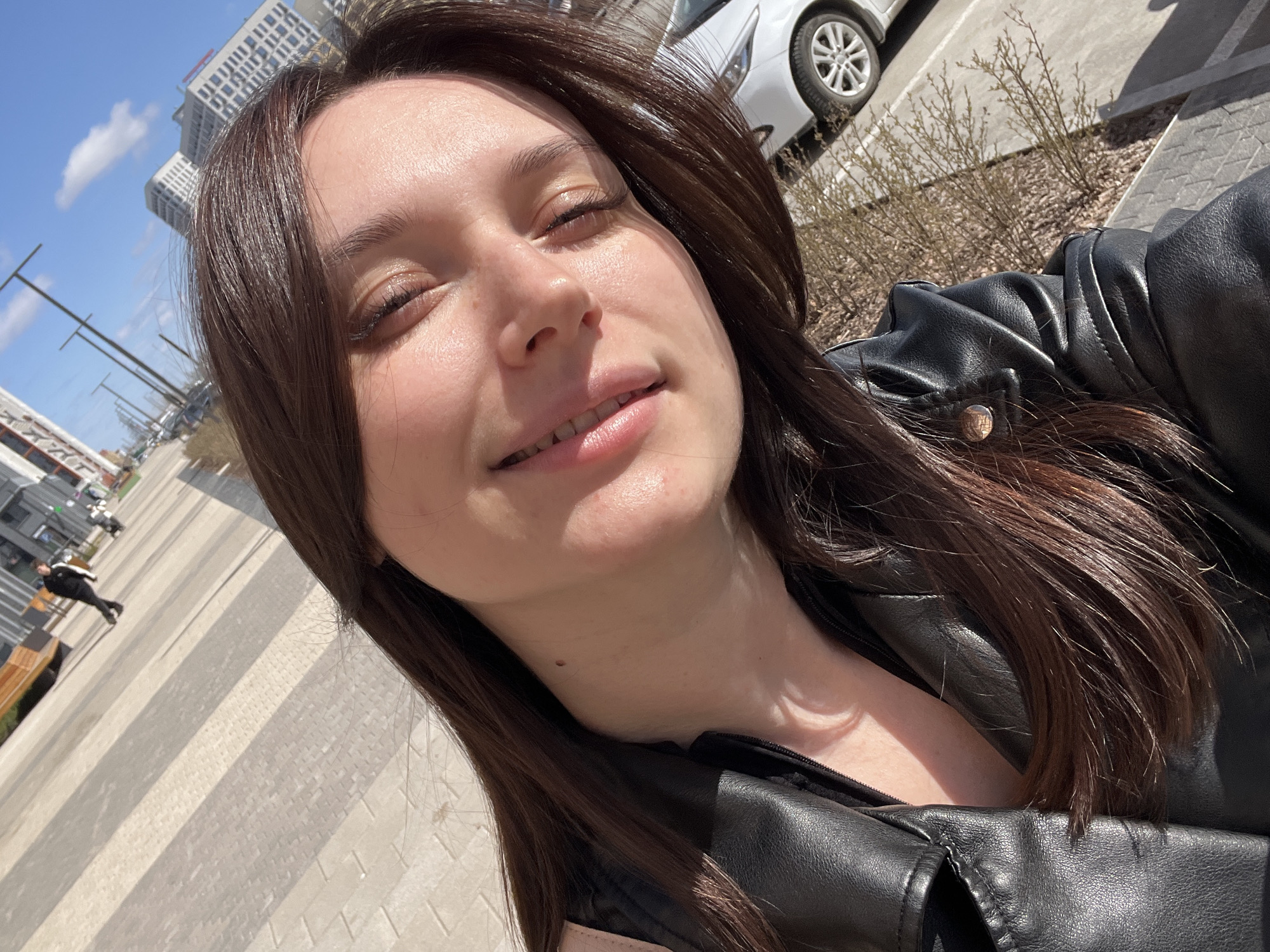 A close-up selfie captures a woman with dark hair, partially closed eyes, and a slight smile. She is wearing a black leather jacket. The background reveals an urban environment with modern buildings, blue sky, and a parked white car. The ground is paved with rectangular stones. A figure is seen in the distance, seemingly sitting or lying on a bench. Based on the architectural style of the buildings and the general layout, the location is likely in the city of Tyumen, Russia.