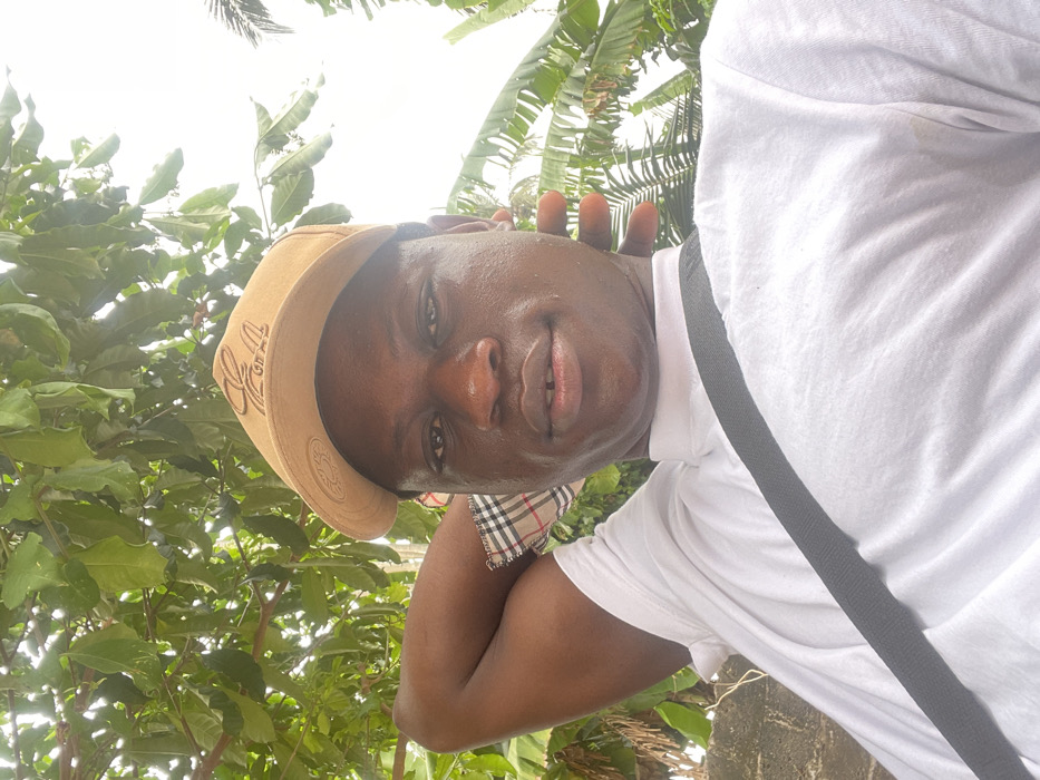 A medium close-up outdoor photograph taken in Ganyingbo, Nigeria, features an adult male with a dark complexion, looking directly at the camera with a neutral expression. He wears a light tan baseball cap with an embossed or embroidered logo on the front, a white t-shirt, and a black strap crossing diagonally from his left shoulder to his right hip. His right arm is bent with his hand positioned behind his head, and a plaid fabric pattern is visible at the inner bicep area beneath the t-shirt sleeve. His left hand is also raised and positioned behind his head. A slight gap is visible between his two upper front teeth. The background consists of dense green foliage, including broad-leafed plants, against an overcast sky visible in the upper right quadrant. A textured light-colored surface, possibly a wall, is partially visible behind his left shoulder.