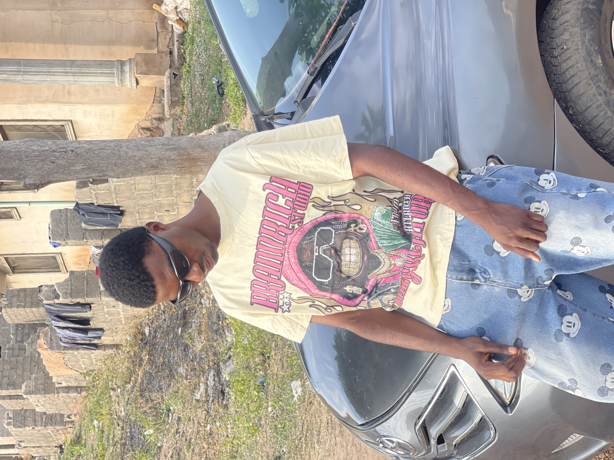 A young man stands outside, leaning against the hood of a dark-colored car. He is wearing sunglasses, a pale yellow t-shirt with a large graphic print, and jeans patterned with Mickey Mouse images. Behind the car, there is a building and some vegetation. The location appears to be Lokoja, Nigeria.