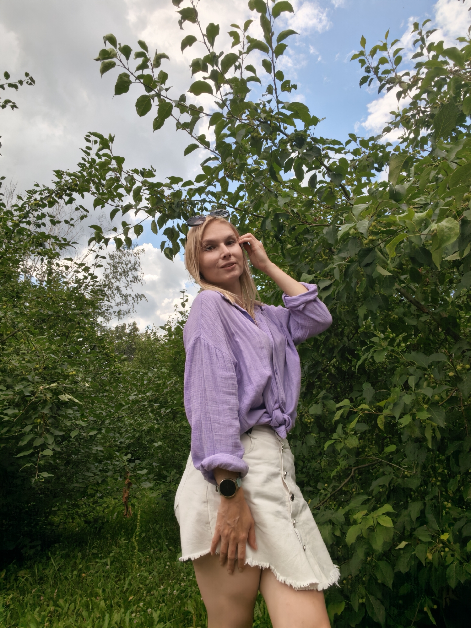 A young woman with blonde hair, wearing a light purple, long-sleeved, button-down shirt tied at the waist and a white denim skirt, stands outdoors amidst lush green foliage. She is looking towards the camera, with her left hand touching her hair. She has a watch on her left wrist. The background consists of dense green trees and a partly cloudy sky. The location is identified as Sterlitamak, Russia.
