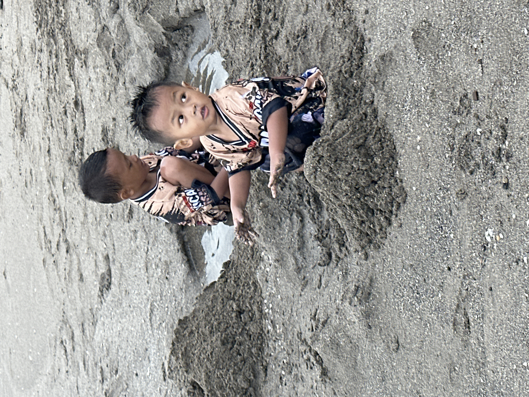 Two young boys are playing in the sand on a beach in North Kuta, Indonesia. The boy in the foreground is wearing a tan shirt with black and white accents and black shorts. He is sitting in the sand with his hands in a pile of sand, looking to his right. The second boy is behind him, to his left, also sitting in the sand. He is wearing a similar tan shirt with black and white accents and black shorts. He is looking down at his hands, which are in the sand. There is a small puddle of water between the two boys. The sand is wet and appears to be mixed with shells. The background is out of focus and appears to be more sand and water.