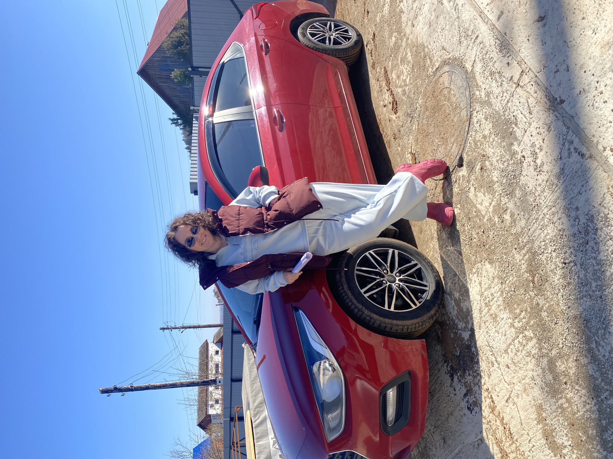 A young woman with sunglasses is casually posing on a red car outside, in the urban/suburban area of Ольховка, Russia. She wears a light blue sweat suit and a brown puffy vest, paired with pink rain boots. She holds a white rolled object, possibly a document or blueprint. The car's tire is partially under her, and she leans against the car's hood. The background includes a clear blue sky, utility poles with wires, houses, and the corner of a building, all suggesting an outdoor, daylight setting within a residential area. The surface on which the car is parked is concrete.