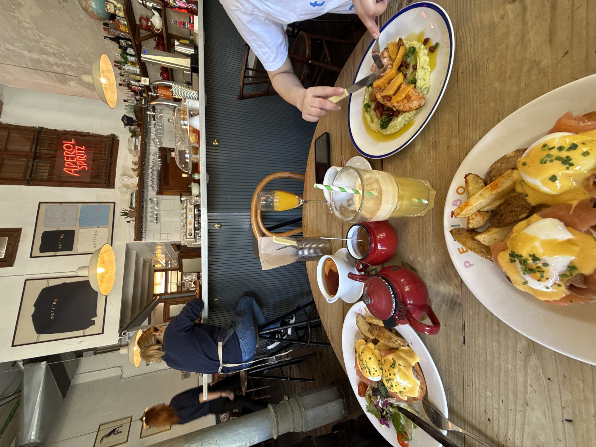 This image shows a table at a restaurant in Barcelona, Spain, cluttered with food and drinks. Two people are visible, one actively eating a meal with a fork and knife. The table holds two plates of what appears to be Eggs Benedict served with potato wedges, and another plate with a different dish featuring salmon. There are also two glasses of orange juice, a blended drink with a straw, a red teapot, and a white teacup. The restaurant's interior is visible in the background, including a bar with bottles and glasses, wall art, and hanging lights. A neon sign reading "Aperol Spritz" is visible. A woman is seen seated at the bar.
