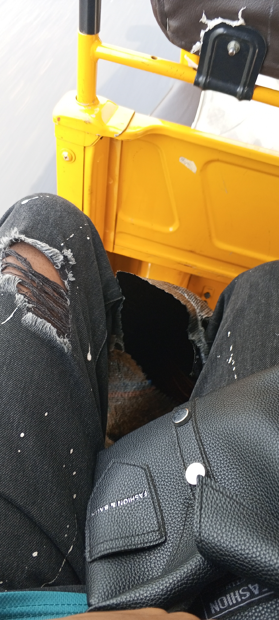 The photo shows a person sitting in a yellow tricycle (Keke Napep) in Uke, Nigeria. The person is wearing ripped black jeans with white paint splatters and has a black textured leather bag resting on their lap. The background includes a portion of the tricycle's yellow metal frame and seat.