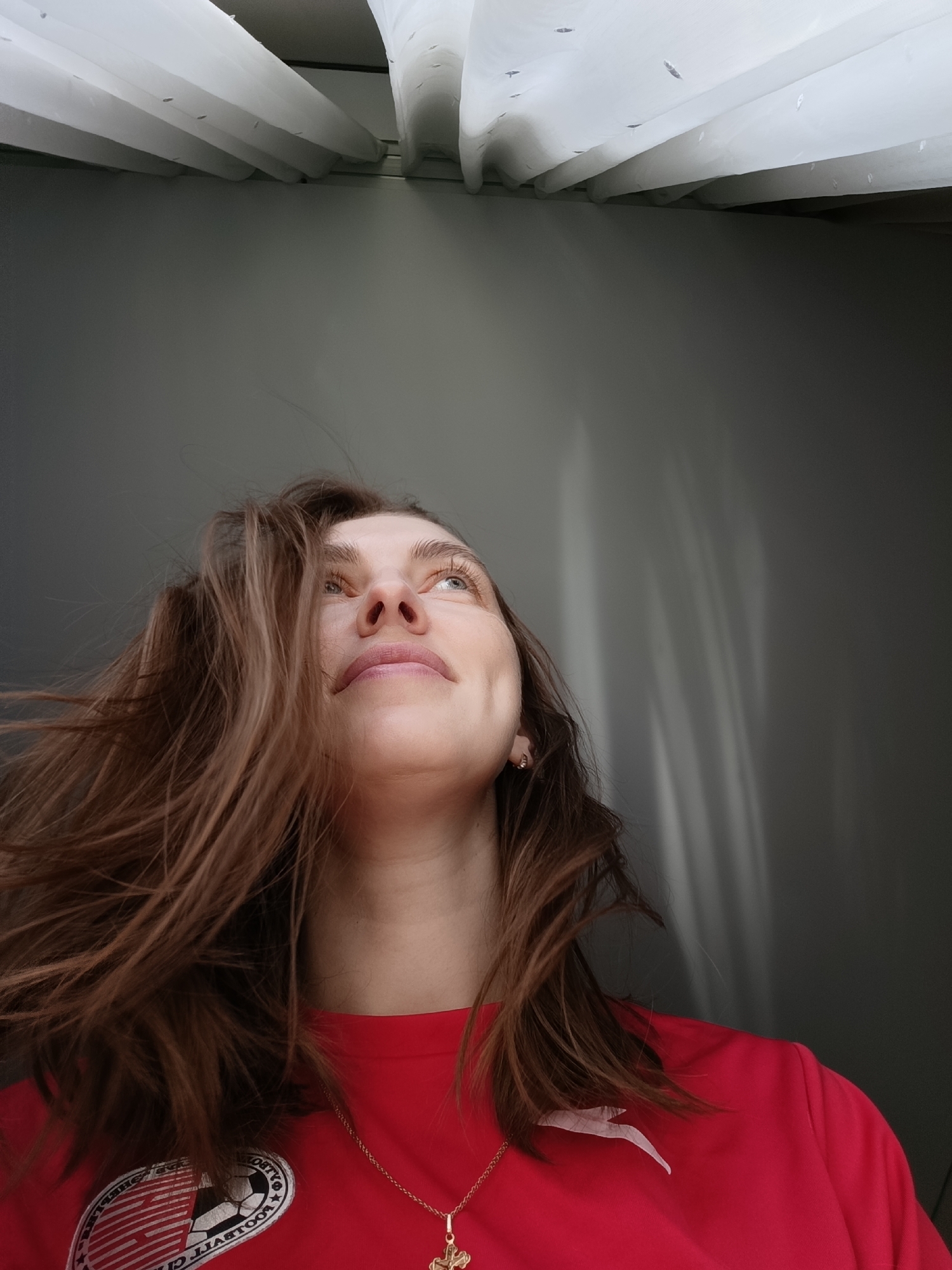 An eye-level indoor portrait captures a woman in Chaikovsky, Russia, looking upwards with a gentle, contemplative expression. Her long, light brown hair, appearing slightly windswept, frames her face. She wears a bright red short-sleeved top with a golden chain and cross pendant around her neck. A small, golden earring is visible in her right ear. On the left side of her chest, a circular logo is present on her shirt. Due to the image's perspective, the text on the logo appears reversed, with partially legible words including "FOOTBALL" and "CHAIKOVSKY" visible. The background features a plain grey wall on the right, illuminated by diagonal patterns of sunlight and shadow. Above her, white, pleated curtains or blinds suggest a window or overhead light source, indicating it is daytime.