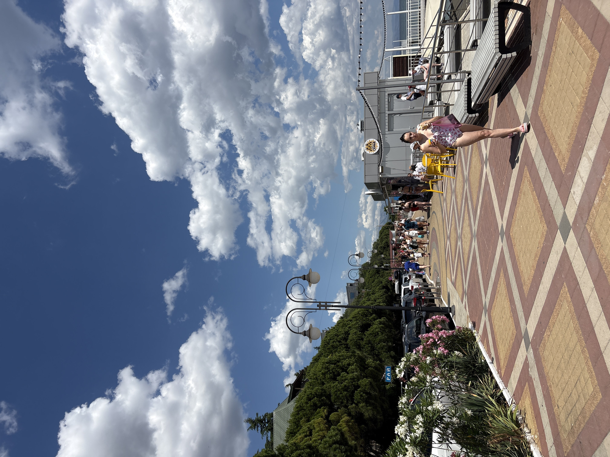 A wide shot captures a sunny day on a boardwalk in Alushta, Ukraine, with a bright blue sky dotted with fluffy white clouds. A woman in a floral swimsuit walks towards the camera, holding an ice cream cone and a shopping bag. To her left, a patterned walkway leads to a bustling promenade with people strolling and seated at yellow tables outside a small cafe. Beyond the promenade, a line of parked cars is visible, bordered by lush green trees. To the right, modern benches are set up along the edge of the boardwalk, with a railing overlooking the sea. The scene conveys a relaxed atmosphere of a summer day at a coastal resort.