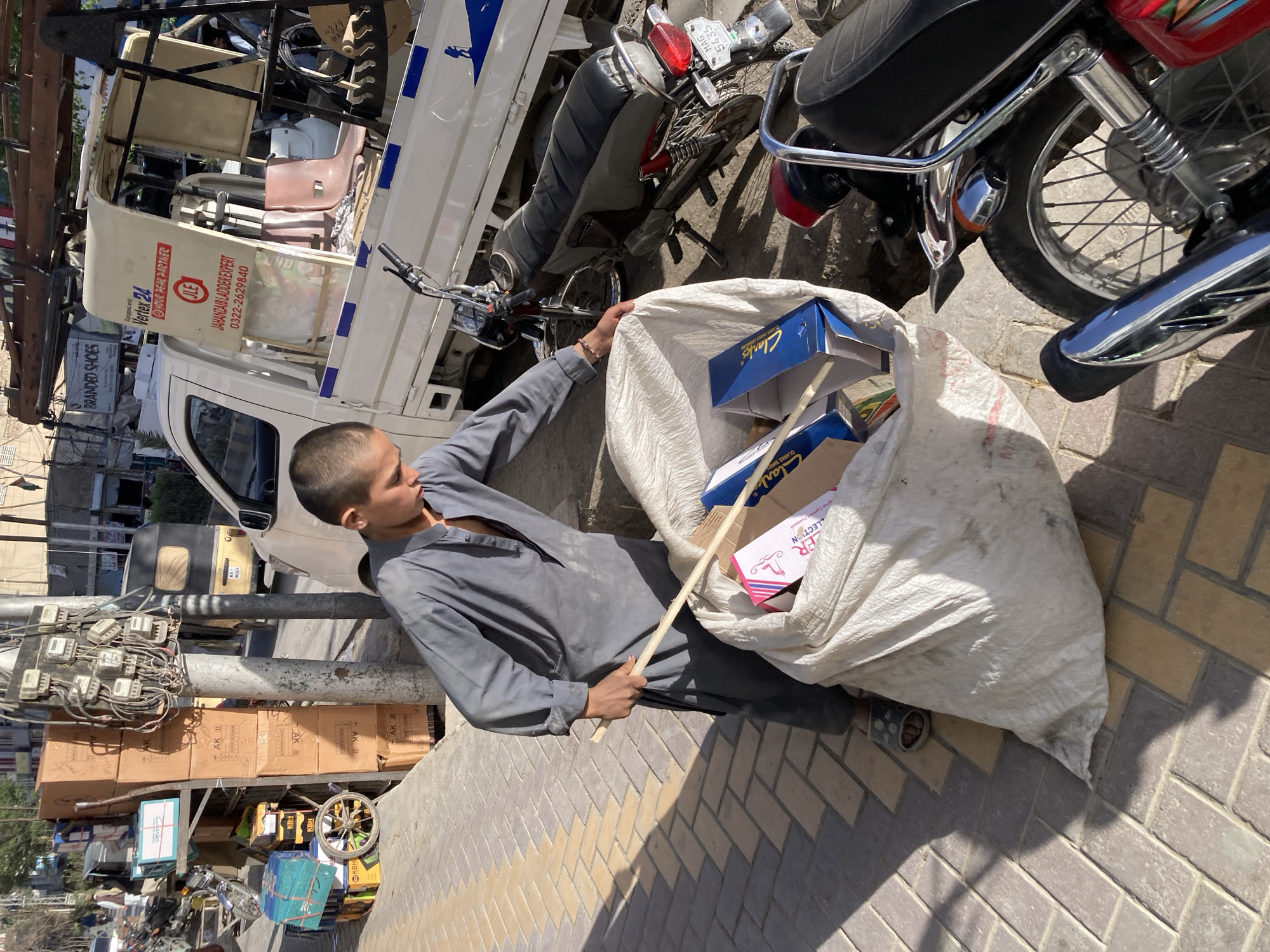 The image, oriented diagonally, captures a street scene in an urban environment, likely a market or commercial area, under bright daylight with strong shadows. The central figure is a young boy, dressed in a grey shalwar kameez and chappals, who appears to be sifting through or carrying a large, white woven sack. He holds a wooden stick, with one end inside the sack.
The sack is filled with various items, notably several blue shoe boxes distinctly labeled "Clarks" and a pink box displaying "ER LECTION." Behind the boy, a white, open-backed pickup truck or motorized rickshaw-like vehicle is visible, laden with goods, including what appears to be ladders. The truck's side bears several advertisements and text: "Vertex 24 @YOUR PEAL PARTNER," "JAHANZAIB LADDER EXPERT," and a phone number "0322-2629840." Another sign on the truck reads "BRANDED SHOES."
Adjacent to the truck, at least two motorcycles are parked, one featuring a license plate with "MAG 5435." To the left, a utility pole is densely packed with multiple electricity meters and intricate wiring. Nearby, stacks of cardboard boxes, some marked "AK II" and "KBH," are piled, possibly for sale or transport, along with a small, wheeled cart. The ground is paved with interlocking bricks. The boy's expression is neutral, focused on his activity, and the vibrant sunlight suggests a clear, warm day. The scene depicts typical daily life and commerce in a South Asian city.