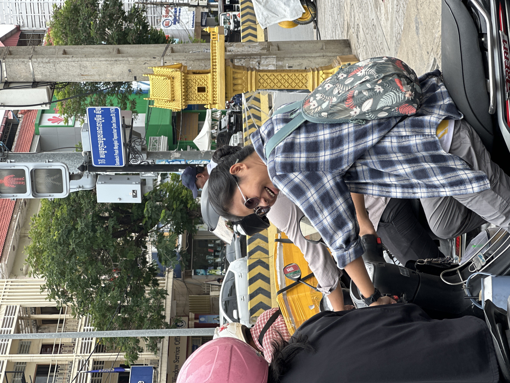 In Khan Boeng Keng Kang, Cambodia, a medium shot captures three individuals on a motorcycle, two women and one person in the rear. The foreground features a woman with sunglasses and a blue and white plaid shirt, her head turned to the left with a wide smile and open mouth. She wears a backpack with a floral print and grey trousers. To her left, another woman, partially visible, wears a pink helmet and a black top. The person in the middle, wearing a red and white plaid shirt, is also partially visible. The motorcycle has a yellow box attached to the back. In the background, a traffic light with red and green pedestrian signals is visible, along with a street sign written in Khmer and English, indicating "Samdech Preah Mongkol Tepeachar Oum Soam St (163)". A golden miniature shrine is mounted on a concrete pole next to the street sign. Buildings, trees, and other vehicles are also present in the urban setting.