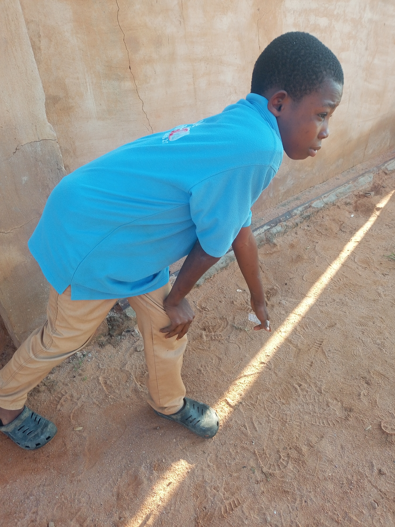 A young boy, appearing to be of African descent, is captured from the side in an outdoor setting. He is wearing a bright blue short-sleeved collared shirt and tan pants. His stance suggests he is bending over or reaching for something on the sandy ground. His left hand is near his left knee, and his right hand is extended downwards, fingers slightly curled, as if touching or picking up an object. He is wearing dark clogs on his feet. The background consists of a textured, light-colored wall, possibly made of stucco or plaster, showing some cracks and imperfections. The ground is composed of loose sand with visible footprints and debris. A diagonal shaft of sunlight cuts across the scene, illuminating a portion of the sand and casting a shadow. The environment appears to be a dry, possibly rural or semi-urban area in Tempure, Nigeria.