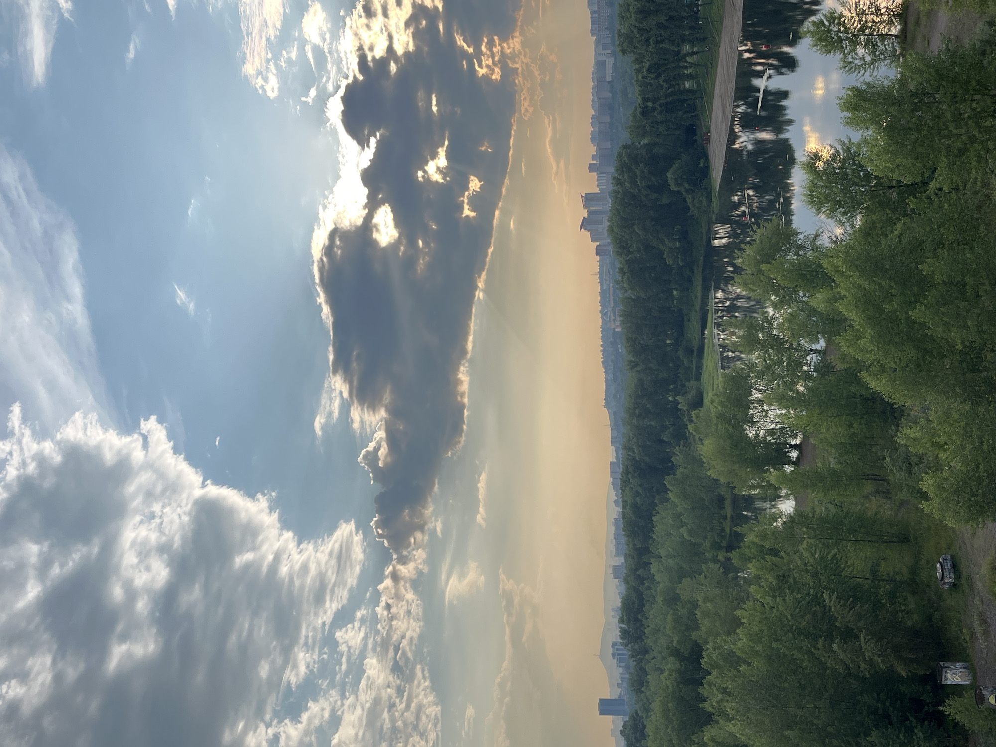 The image captures a panoramic view of a river flowing through a city, likely Krasnoyarsk, Russia, under a dramatic sky. The foreground is dominated by lush green trees that line the riverbanks, with a small section of a sandy beach visible on the right side of the river. The water reflects the sky and the surrounding foliage, creating a serene atmosphere. In the distance, a city skyline with various buildings and a prominent skyscraper is visible, set against a hazy horizon. The sky is filled with a mix of light and dark clouds, with sunlight breaking through in some areas, casting rays of light. The overall impression is that of a late afternoon or early evening scene, with the natural beauty of the river and trees juxtaposed with the urban landscape.