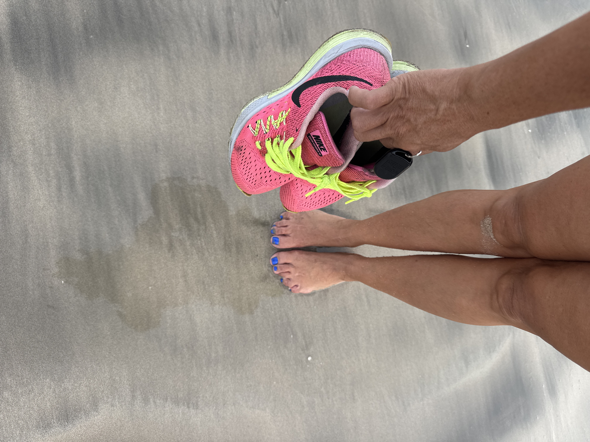 The image depicts a person standing barefoot on a sandy beach. The sand appears slightly damp and smooth. The person is holding a pair of pink Nike running shoes with yellow laces in their right hand. The person has tan legs and blue nail polish on their toenails. The image does not contain any clear features to indicate it is in Jimbaran, Indonesia.