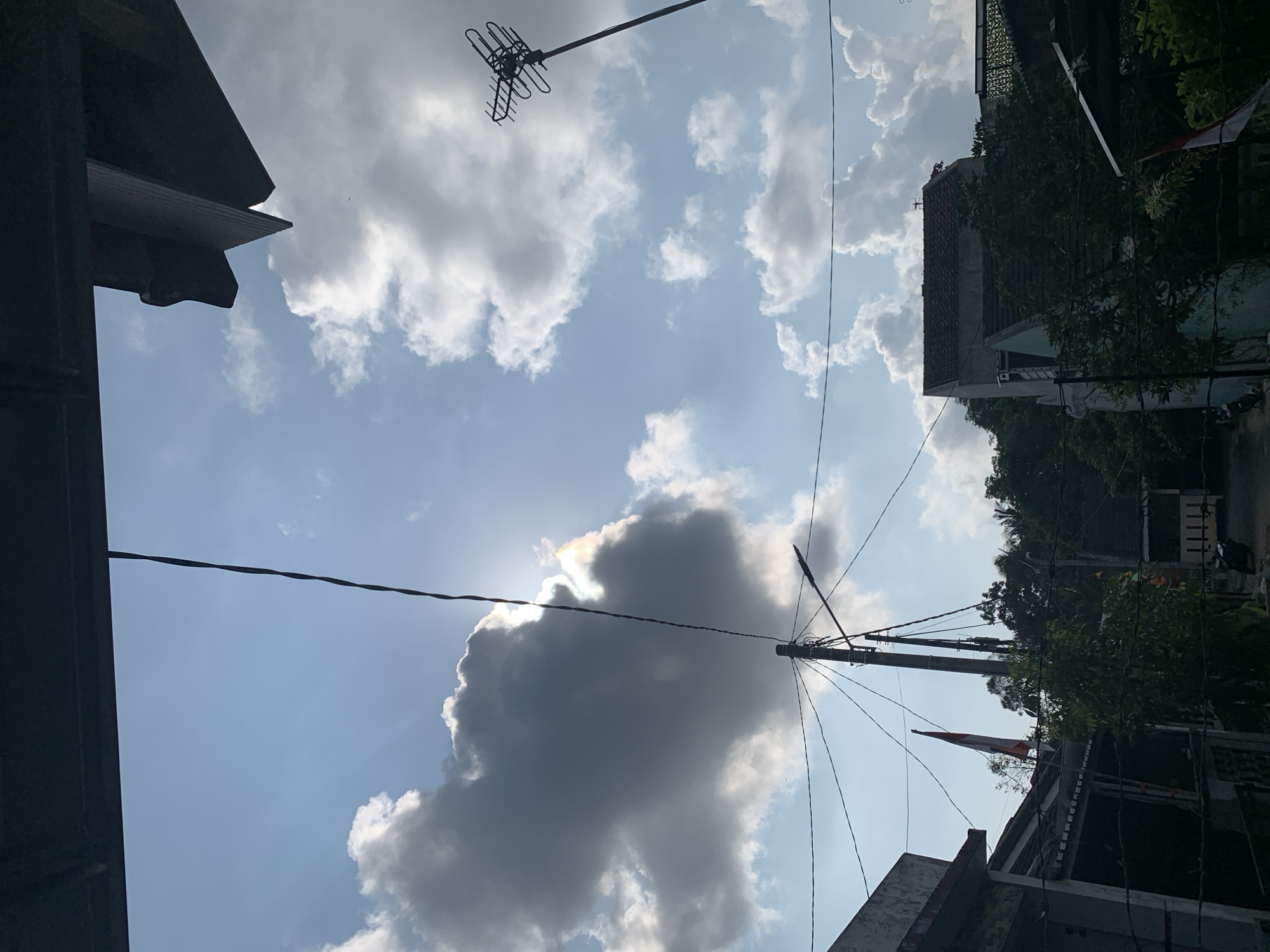 The image depicts an outdoor scene, likely in a residential area of Utama, Indonesia, viewed from beneath an overhanging structure. The sky is a prominent feature, with a mix of blue sky and dramatic, dark gray clouds partially obscuring a bright sun. A thin, dark electrical wire stretches diagonally across the frame, originating from the top left and disappearing towards the lower right. To the left, a utility pole with multiple wires and a streetlight is visible. Behind it, a large, dark cloud dominates the left side of the sky, with the sun peeking through its edge, creating a halo effect.

In the foreground and midground, several elements suggest a human environment. On the left, part of a building with a red-tiled roof is visible, along with a tall, slender pole displaying a flag that appears to be the Indonesian flag due to its red and white colors. Further to the right, another utility pole with an antenna on top stands against the sky. Below the sky and between the utility poles, dense green foliage and trees are visible, interspersed with the roofs of houses. Some of these houses have red-tiled roofs, while others have lighter-colored walls and roofs. A glimpse of a motorcycle can be seen on the ground in the lower right portion of the image. The overall impression is of a slightly overcast, yet bright, day in a built-up area with a clear Indonesian presence.