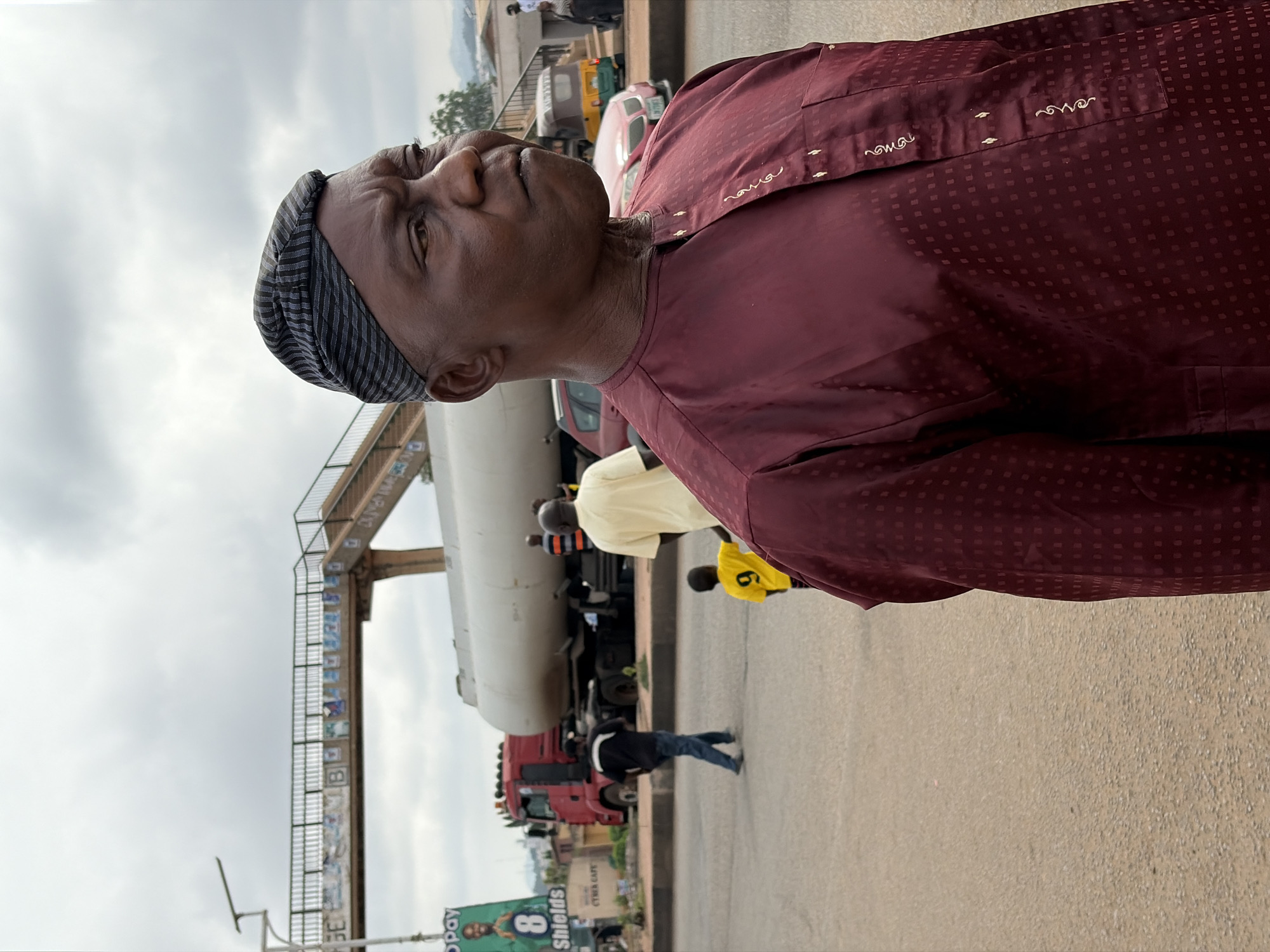 The image displays an outdoor street scene under an overcast sky, likely during the daytime. The primary subject is a man of African descent, positioned in the mid-foreground, looking slightly upwards and to his left. He wears a black and grey striped kufi cap and a maroon-colored, long-sleeved traditional top featuring a subtle square pattern and embroidered accents around the neckline. His expression appears neutral or contemplative.
Behind him, a bustling street unfolds with a prominent white tanker truck, distinguished by its red cab, traversing the road. Several other individuals are visible in the midground; one person wears a light yellow shirt, another dons a bright yellow shirt with the number '6' on the back, and a third, dressed in a black jacket and blue jeans, walks away from the viewer. Other vehicles, including a red car and possibly a tricycle, are scattered along the road.
To the left, a large pedestrian bridge spans the roadway, featuring metal railings and concrete supports, with some faint posters or graffiti adhered to its structure. In the far distance, low hills are visible beneath the cloudy sky.
In the bottom-left corner, a partial green billboard is discernible, displaying text such as "Opay" and "Shields," along with the number "8" and a faint image of a person. A smaller sign beneath it also hints at "CYBER CAFE." The overall setting suggests a common street environment in an urban or semi-urban area, consistent with Gura Top, Nigeria.