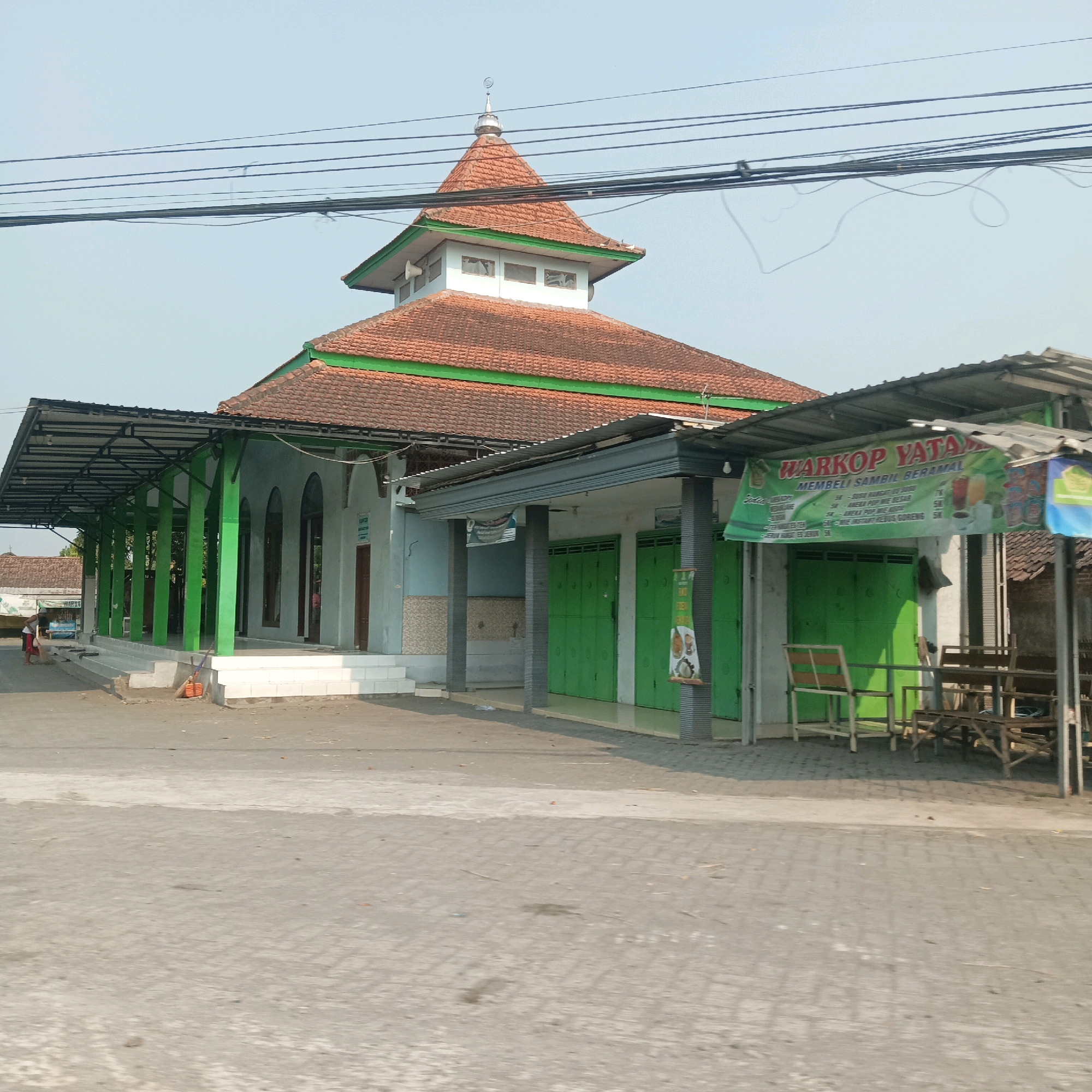 The image captures a street scene in Kayen, Indonesia, featuring a mosque and adjacent commercial establishments. The mosque, with its distinctive tiered roof and minaret topped with a crescent moon, is the central structure. A covered walkway supported by green pillars extends along the front of the mosque. To the right of the mosque, a "WARKOP YATANA" sign is prominently displayed above green shutters, indicating a coffee shop or food stall. This stall appears to be selling various beverages, with images of drinks and food items visible on a smaller sign. Wooden tables and chairs are set up outside the stall, suggesting an outdoor seating area. The ground is paved with interlocking blocks, and a street runs in front of the buildings. A person is seen sweeping the area near the mosque's entrance. The sky is clear and bright, indicating daylight. The overall scene depicts a functional, everyday corner of the town, blending religious and commercial activities.