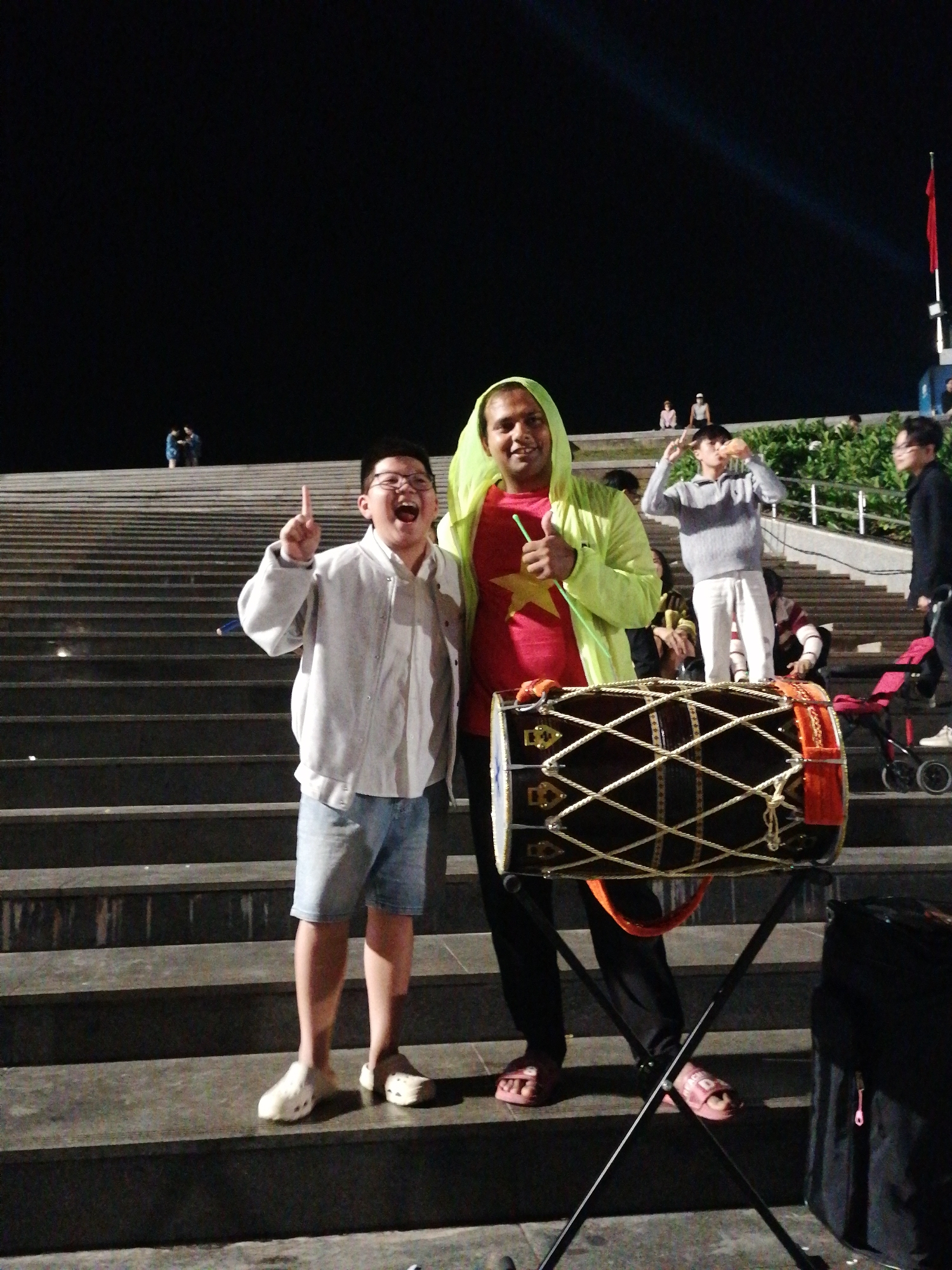 In Da Lat, Vietnam, at night, two young men are posing for a picture on a wide set of outdoor stairs. The man on the left, a child, is wearing a light gray button-down shirt and denim shorts. He is smiling widely and pointing upwards with his right index finger, wearing light-colored slip-on shoes. The man on the right is wearing a bright yellow hooded jacket over a red shirt with a yellow star. He is holding a green stick and gives a thumbs-up gesture. He is wearing dark pants and pink slide sandals.
In front of them, a large drum is set up on a stand. The drum is dark with ornate gold accents and is strung with white rope. Behind them, other people are visible on the stairs and in the background. Some are seated, while others are standing. A red flag is visible on a flagpole in the distance, and a spotlight beam cuts through the dark sky. The scene suggests a public gathering or performance. The lighting indicates it is nighttime, and the presence of a spotlight suggests an organized event.