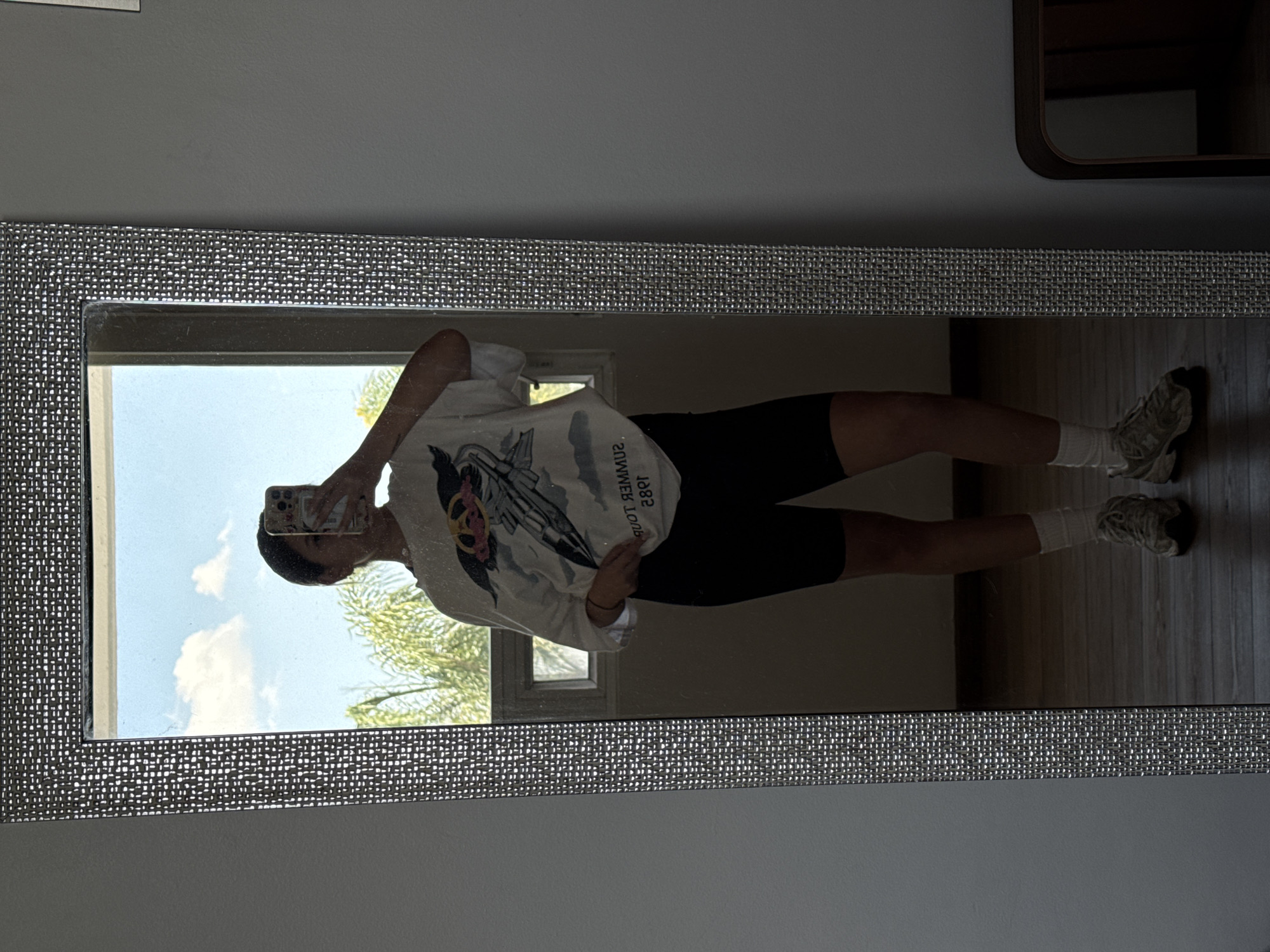 A person is reflected in a mirror, holding a cell phone up to their face. They are wearing a white t-shirt with a graphic print, black shorts, and white socks with sneakers. The mirror reflects a view of a clear blue sky with fluffy white clouds and the foliage of a tree, likely from a window within the room. The location appears to be Heroica Puebla de Zaragoza, Mexico based on the architectural style and general climate that is seen through the window. The mirror has a decorative woven-style frame. It's placed against a light gray wall. Adjacent to the mirror on the right is a glimpse of a dark brown wooden structure, likely a shelf or cabinet. The floor is made of wooden planks.