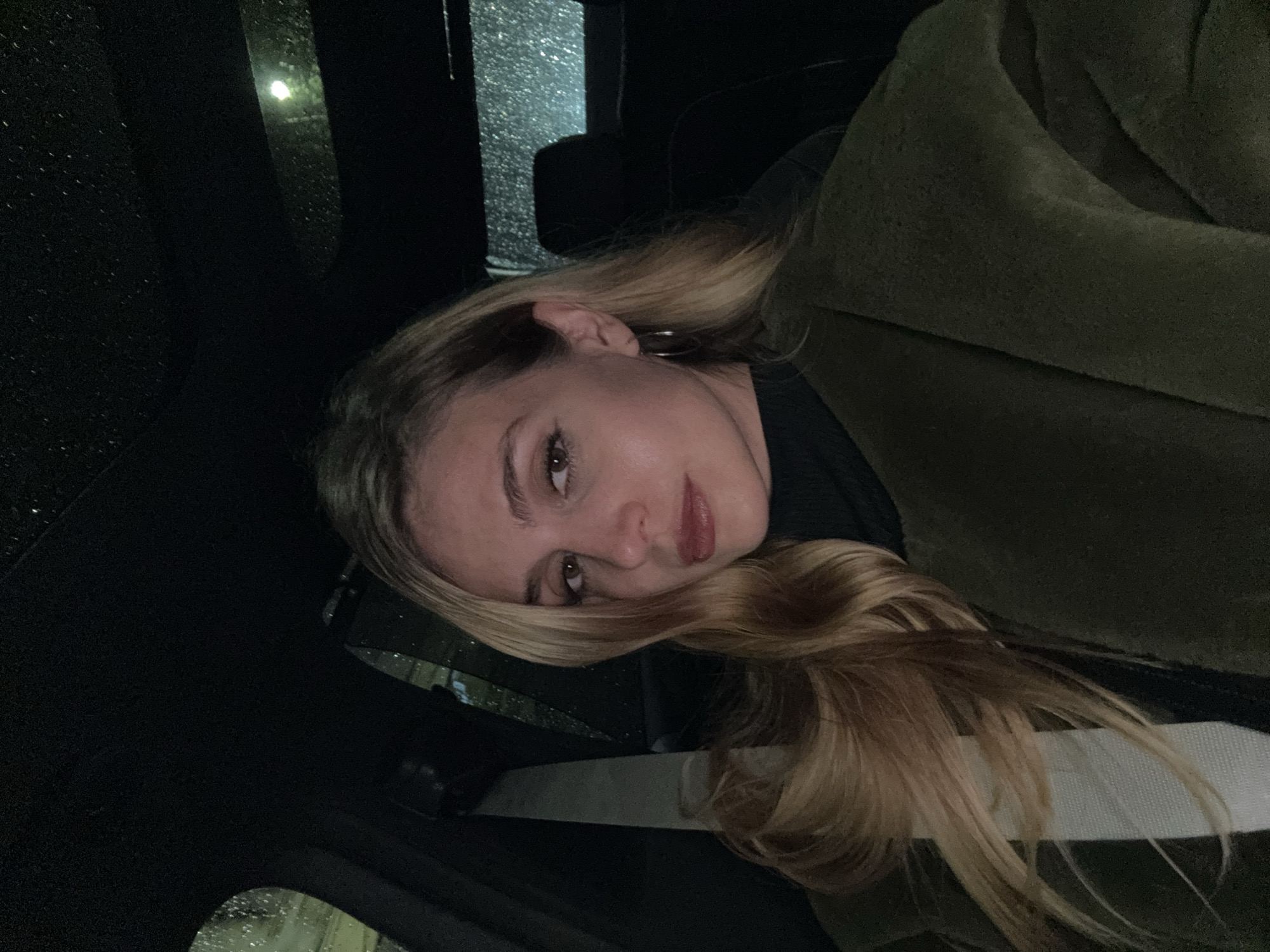 A young woman with long, wavy blonde hair is captured in a medium close-up shot, appearing to be inside a car in Tbilisi, Georgia. She is looking directly at the camera with a neutral, slightly contemplative expression, her lips tinted with a reddish hue. She wears a dark top and an olive green coat, with a white seatbelt visible across her chest. The car's interior is dark, suggesting the photo was taken at night or in very low light. Through the car window on the left side of the frame, numerous raindrops are visible, indicating it is raining outside. Faint glints of light, possibly from streetlights, reflect off the wet glass. The overall scene conveys a dim, intimate atmosphere within the vehicle during inclement weather. No visible text is present in the image.