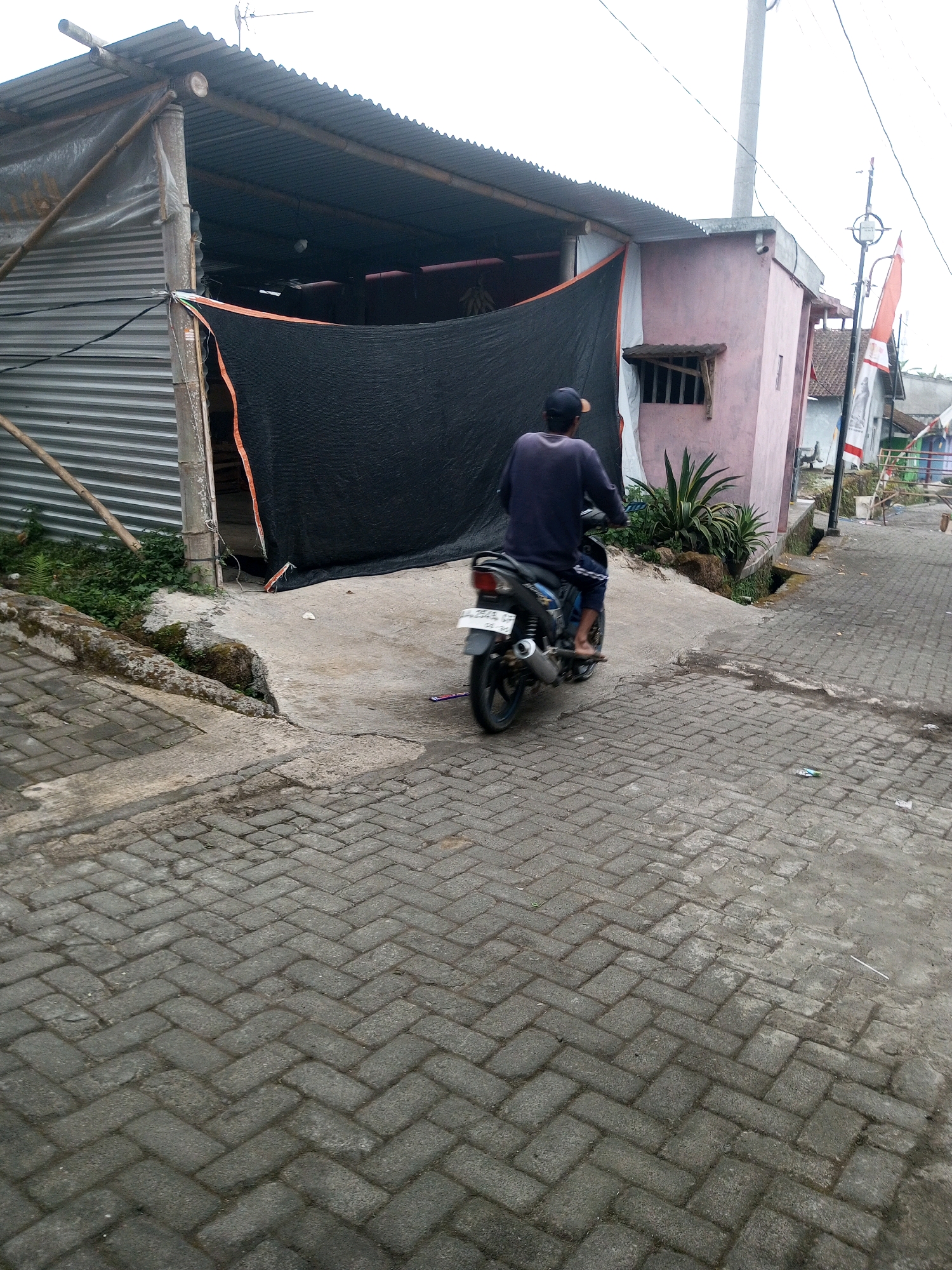 In Andongsili, Indonesia, a person on a motorcycle is seen from behind, heading away from the viewer. The person is wearing a dark long-sleeved shirt, dark pants, and a dark baseball cap. The motorcycle is a dark color with a license plate that is partially visible. The scene is set on a brick-paved street with a concrete pathway leading to a building. The building appears to be a shop or a workshop, with a corrugated metal roof supported by bamboo poles and a large black tarp covering the entrance. Next to the building, a pink-colored structure with a small, barred window is visible. Green vegetation, including some ferns and a spiky plant, grows along the side of the pathway and the building. A street lamp with a flag attached to it stands on the right side of the image. The sky is overcast.