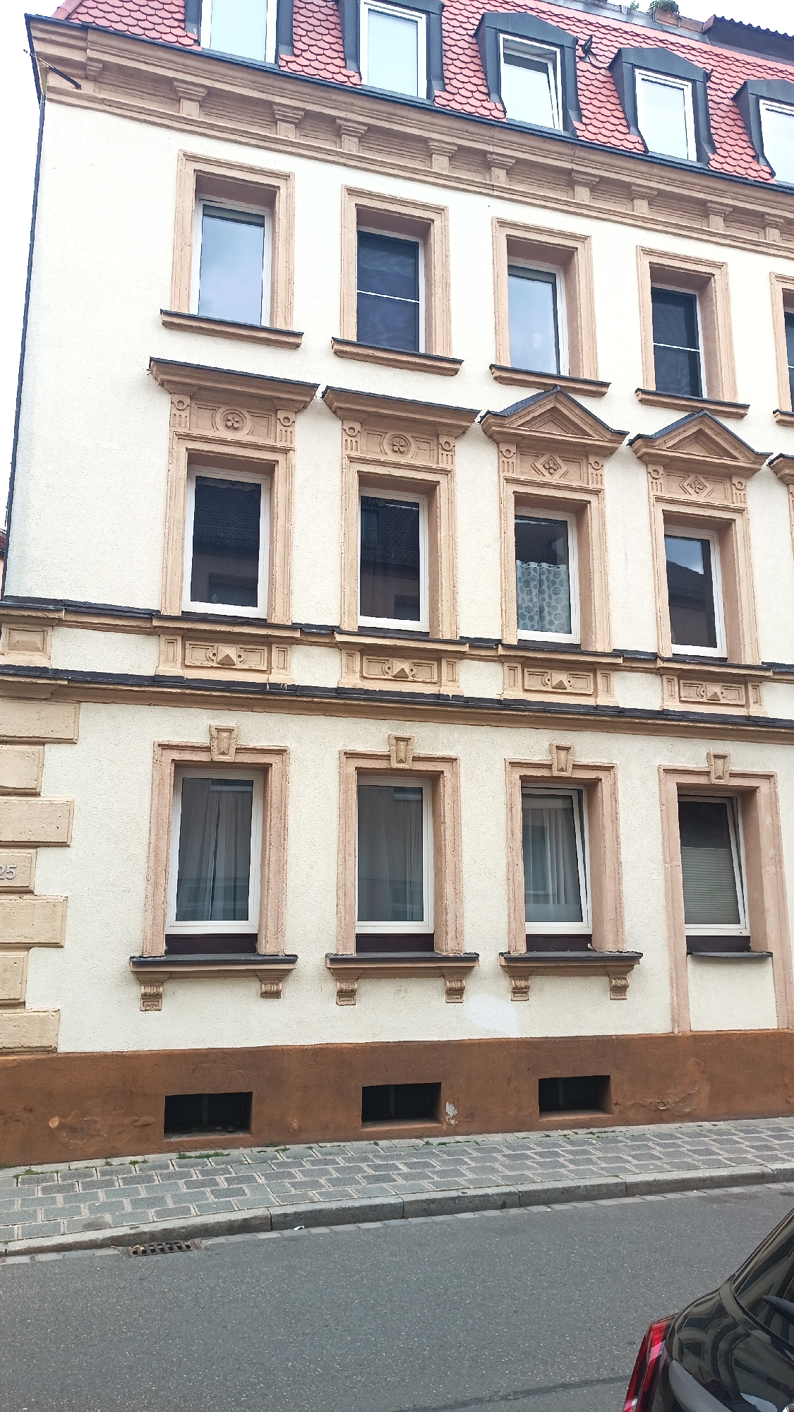 The image shows the facade of an apartment building in Nuremberg, Germany. The building has multiple stories with several windows on each floor, decorated with ornate beige trim. The roof is covered in red tiles and features dormer windows. The lower part of the building has a brown base, and below that, a sidewalk made of grey cobblestones is visible, bordering a street. The building appears to be a residential structure.
