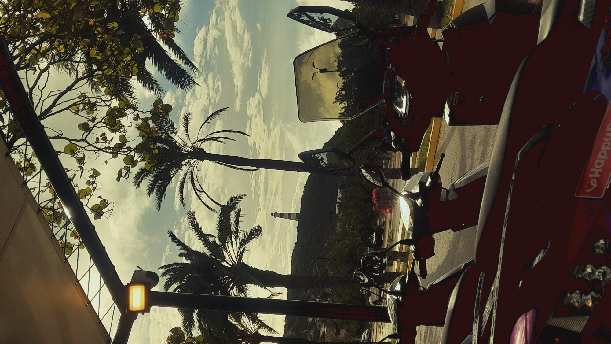 A scene captures a row of parked red scooters under a canopy, with palm trees and a lighthouse in the background. The location is identified as Pedro González, Venezuela. In the foreground, the handlebars and front sections of the scooters are visible, with one scooter displaying a "Happy Bike" sticker. The scooters are parked on an asphalt surface, with a yellow curb separating it from a patch of grass and trees.
Behind the scooters, palm trees frame a view of a verdant hill. At the crest of the hill stands a tall lighthouse with red and white stripes. The sky above is cloudy, with patches of sunlight breaking through. A street light is visible mounted on a pole near the scooters. The overall atmosphere suggests a sunny day in a tropical or subtropical region.