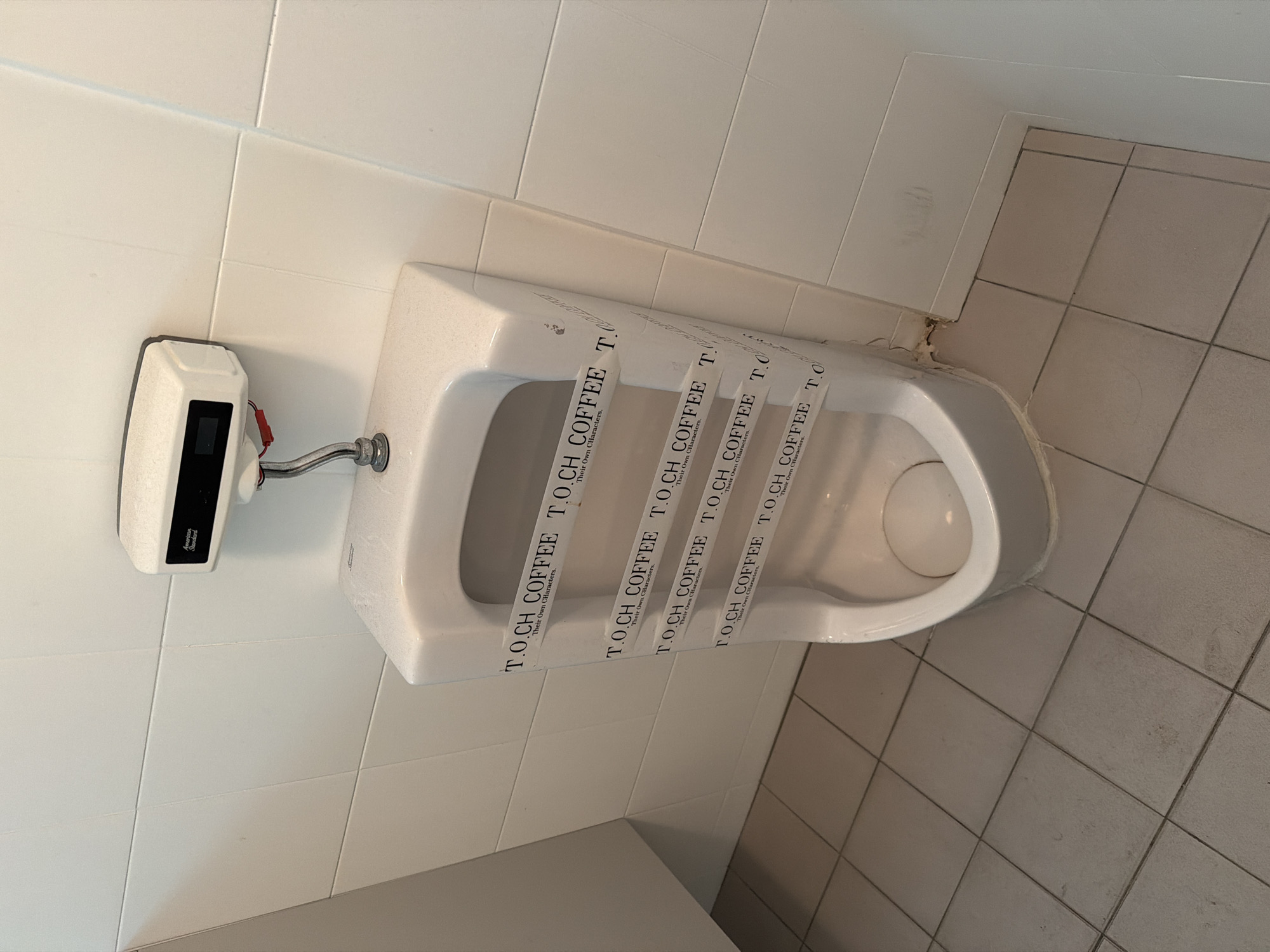 This image shows a urinal with white ceramic tiles and a floor covered in square tiles. The urinal is partially covered with strips of tape that say "T.O.CH COFFEE Their Own Characters" repeatedly. The flushing mechanism is visible on the left side, mounted on the wall. The specific location in Seoul, South Korea, cannot be determined from the given image.