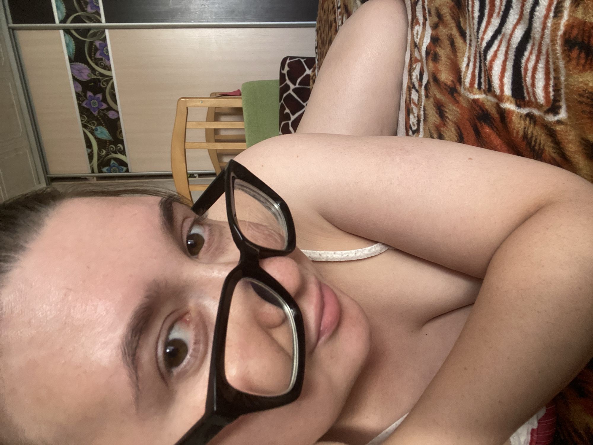 A young woman wearing black-framed glasses and a white strap top is lying on a bed with a tiger-striped blanket. She is looking directly at the camera, with her head tilted. Her face is close to the lens, with her right eye and part of her left eye visible. The background shows a closet with floral patterned doors and a wooden chair with a green cushion. The location is a bedroom in a house in Chaikovsky, Russia.