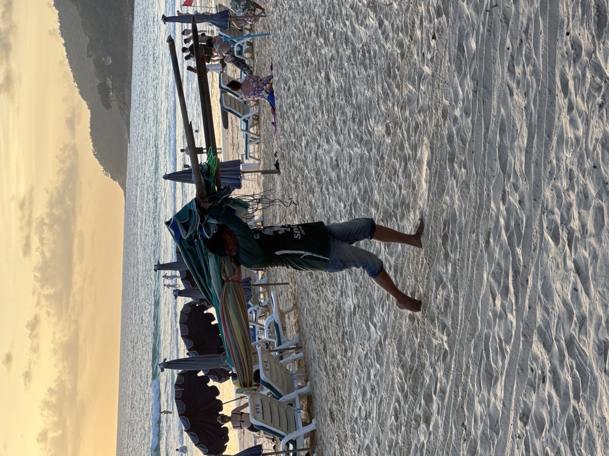 A person is carrying a folded beach umbrella on their shoulder at Karon Beach, Thailand. The beach is sandy with several rows of lounge chairs and umbrellas visible. In the background, the ocean meets the horizon under a sky filled with soft clouds, illuminated by the setting sun. There are other people on the beach, some wading in the water, others sitting on lounge chairs or towels. The overall scene depicts a typical evening on a popular tourist beach.