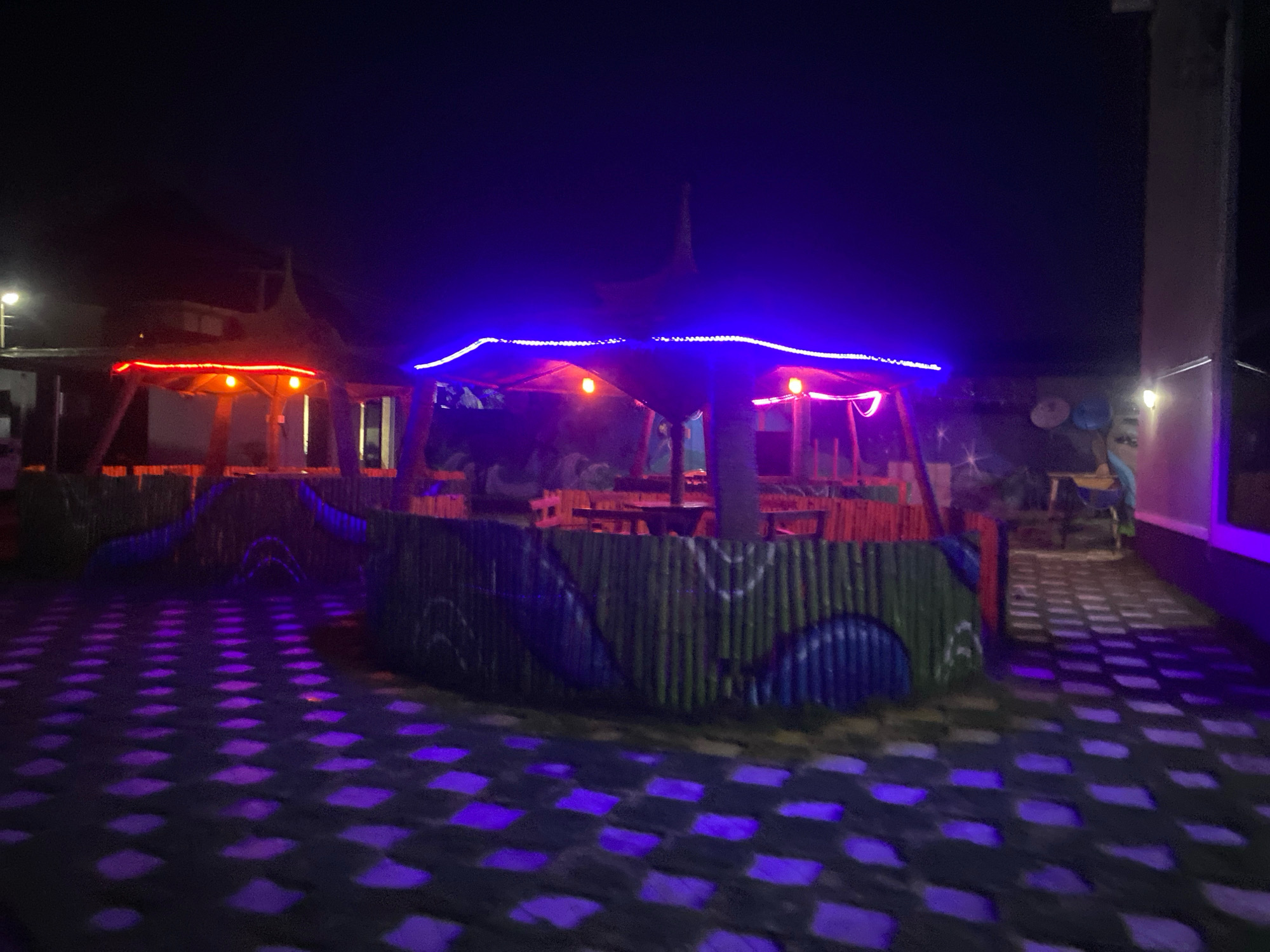 The image shows an outdoor seating area at night, illuminated by colorful lights. The seating area appears to be a restaurant or a bar, with tables and chairs visible. The ground is paved with square tiles, some of which are lit with purple lights. There are bamboo fences around the seating area, decorated with painted designs. In the background, there are more lights and structures, suggesting a lively atmosphere. The overall impression is one of a vibrant and festive nighttime venue.