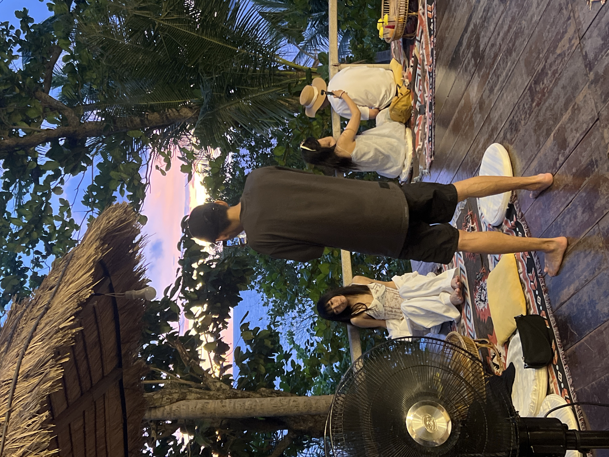 In Karon, Thailand, a group of four individuals are positioned on a wooden deck overlooking the sea at dusk. A large black fan is situated on the left side of the frame. In the center, a person wearing black shorts and a dark t-shirt stands with their back to the camera. To their left, a woman with long black hair, dressed in white, sits on the deck, her legs crossed. To the right of the standing person, a woman in a white dress and a man wearing a light-colored hat are seated. The woman in the white dress is reaching towards the man. The background displays a serene ocean scene under a twilight sky with hues of pink and orange. The deck is adorned with patterned rugs and cushions.