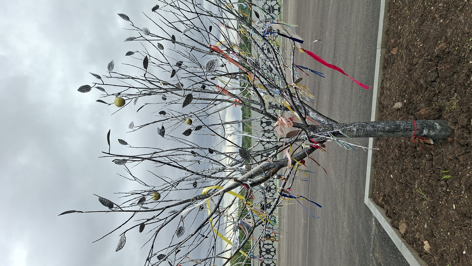 An artificial tree with gray metal leaves and a gray trunk decorated with colorful ribbons stands in a planted area in the city of Nytva, Russia. The tree has two yellow apple-like objects and is planted in a bed of dark brown soil. The tree is located next to a paved area and a decorative metal fence with a body of water visible in the background. The sky is overcast and gray.