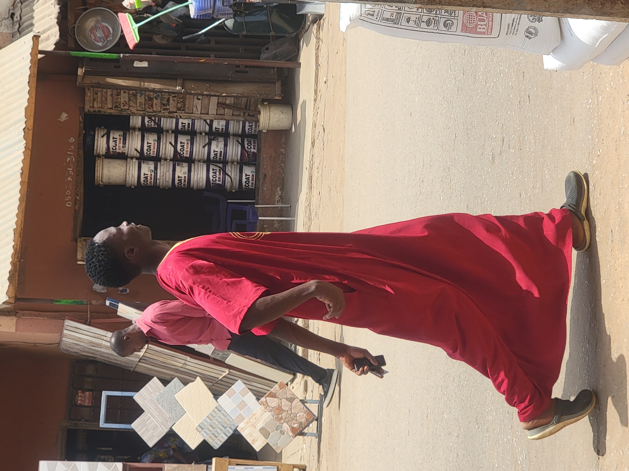 Here's a detailed description of the image:
**Location:** The image appears to be taken in a commercial area, likely in Maiburiji, Nigeria, based on the presence of building materials and consumer goods.
**People:**
* A man wearing a red robe is the main subject. He appears to be walking, holding what seems to be a mobile phone.
* Another man is partially visible behind him, wearing a pink shirt and darker pants.
**Objects:**
* Stacks of paint cans are visible in the background, labeled "ECOAT".
* A display of ceramic tiles is in the foreground.
* A white bag labeled "BUA GYPSUM PLAS" is partially visible in the upper right corner.
**Environment:**
* The setting appears to be an outdoor area, possibly a street or sidewalk.
* The lighting is bright and suggests it is daytime.
* The overall impression is a bustling commercial area with various shops or stalls selling building materials.