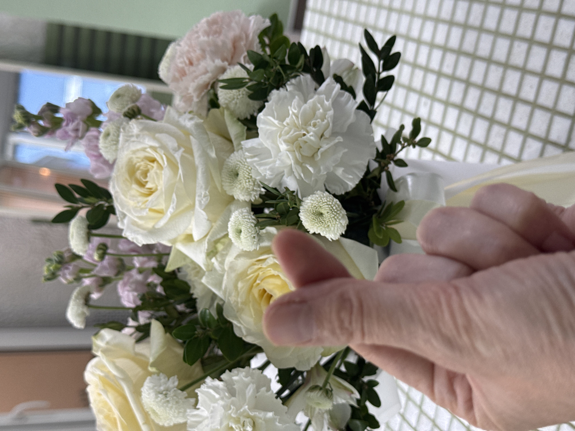 A close-up photograph shows a bouquet of white and pastel flowers held in front of a grid-patterned surface. A person's hand is positioned in the foreground, making a "heart" gesture with their thumb and forefinger. The bouquet includes white roses, white carnations, and small white chrysanthemums, along with some greenery and lighter purple sprigs of flowers. The flowers are tied with a white ribbon. A window is visible in the upper left of the frame. The location in Izhevsk, Russia, is not discernible from the information provided in the image.