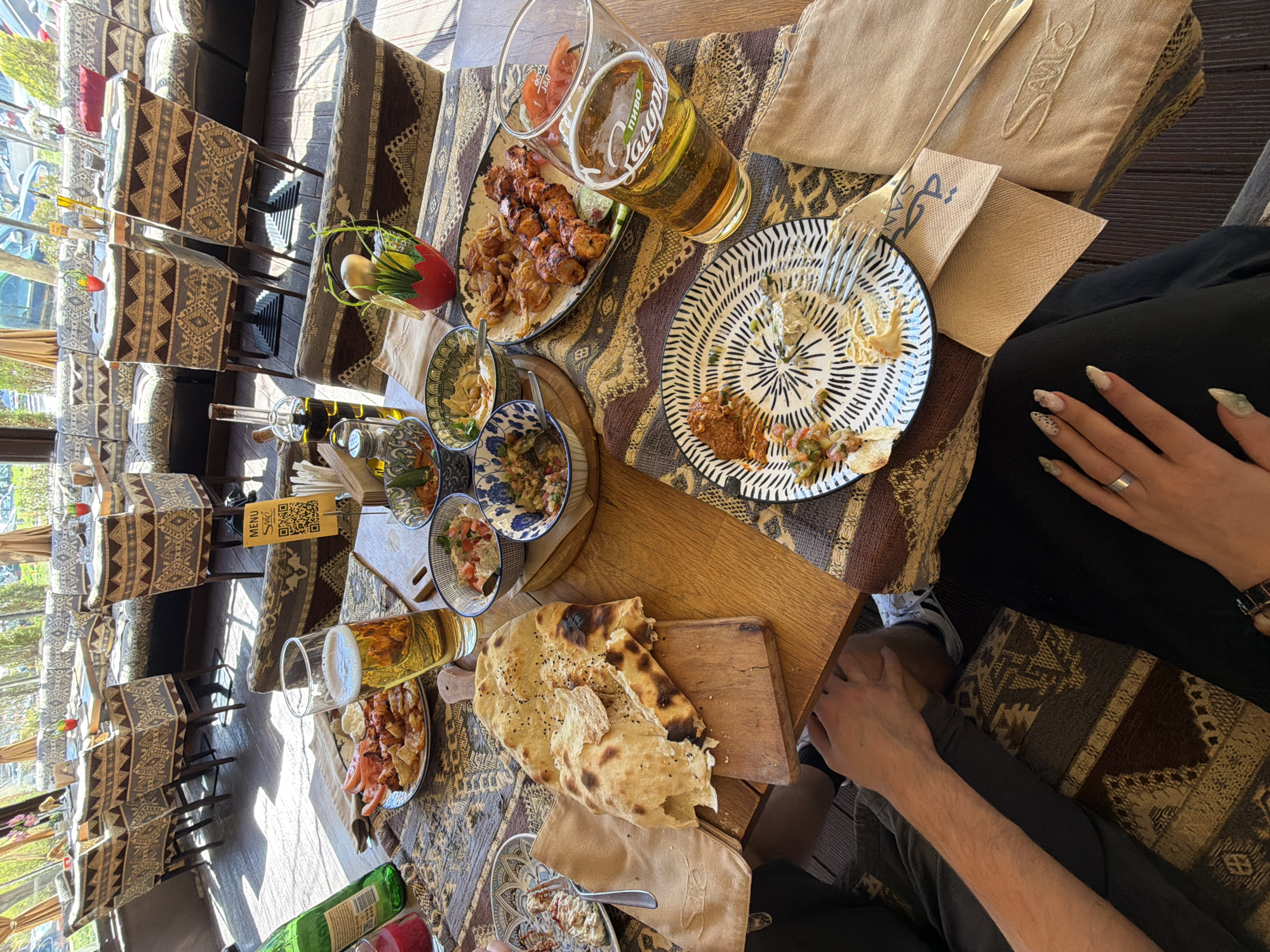 A high-angle shot depicts a table laden with food and drinks in what appears to be a restaurant in an unknown location. The table is wooden and partially covered by a patterned textile. Plates containing remnants of food, skewers of meat, and small bowls filled with various dips and salads are scattered across the table. A tall glass of beer sits near one plate, and a flatbread rests on a small wooden cutting board. Cutlery and napkins are also present. The hands of two individuals are visible, one with manicured nails adorned with patterns and a ring on one finger. Seating with patterned upholstery lines the restaurant's interior, which is brightly lit by natural light coming through large windows. Outside, trees and other buildings can be seen. The overall setting suggests a shared meal in a restaurant environment.