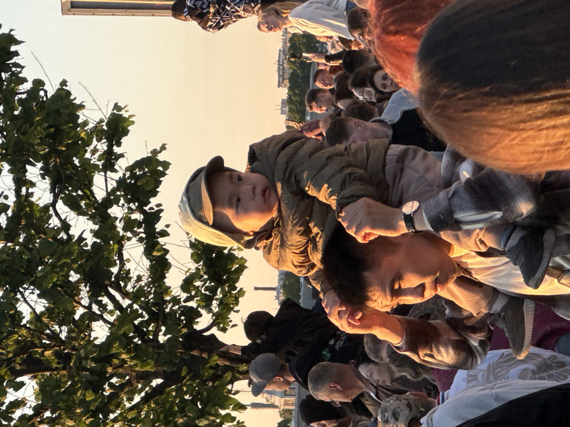 A group of people are gathered outdoors, with some seated on the ground and others standing. The focus is on a child being held by an adult, both looking towards the camera. The child is wearing a green jacket and a cap, while the adult has on a checkered shirt and a watch. In the background, more people are visible, along with trees and buildings that suggest an urban setting, possibly near a waterfront given the hints of structures consistent with Saint Petersburg. The lighting indicates it is late afternoon or early evening, with warm, golden sunlight illuminating the scene. The overall mood appears relaxed and communal, with people appearing to be enjoying a shared moment outdoors. There is no visible text in the image.