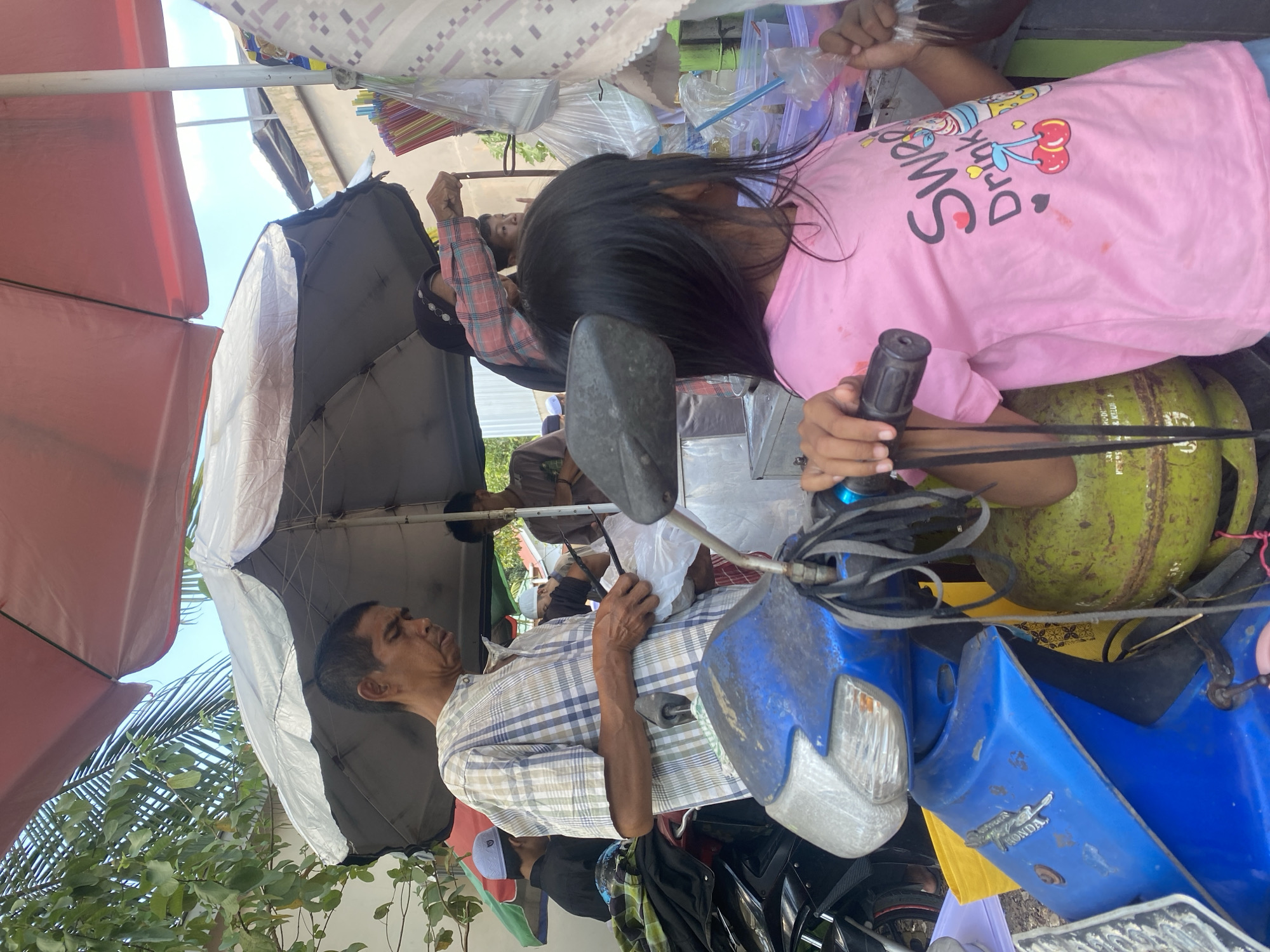 In Banjarbaru, Indonesia, an outdoor scene unfolds at a street-side vendor stall. A man in a checkered shirt is seen holding plastic bags and what appear to be cooking utensils, possibly preparing food or drinks. He is standing near a blue motorcycle, which has a green gas tank attached to its frame. To the right, a young girl with long black hair, wearing a pink t-shirt with the text "Sweet Drink" and cartoon cherries, is holding a plastic bag of a dark liquid. Behind the girl, another woman in a pink plaid shirt is reaching towards the vendor. Several other individuals are partially visible in the background, suggesting a busy market or roadside establishment. The stall is shaded by two large umbrellas, one red and one silver, and there are shelves with various items, including colorful drinking straws. The environment is typical of a street vendor setting in Southeast Asia, with vegetation and buildings visible in the background under a bright sky.